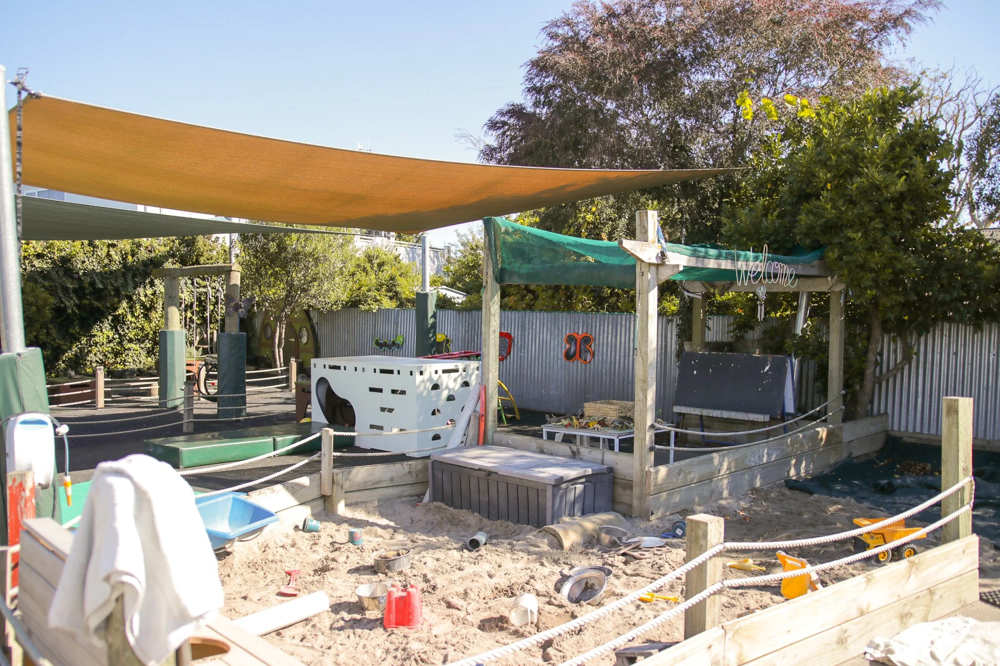 A playground area under construction with sand, toys, and various equipment, surrounded by a fenced yard with trees and shade sails overhead.