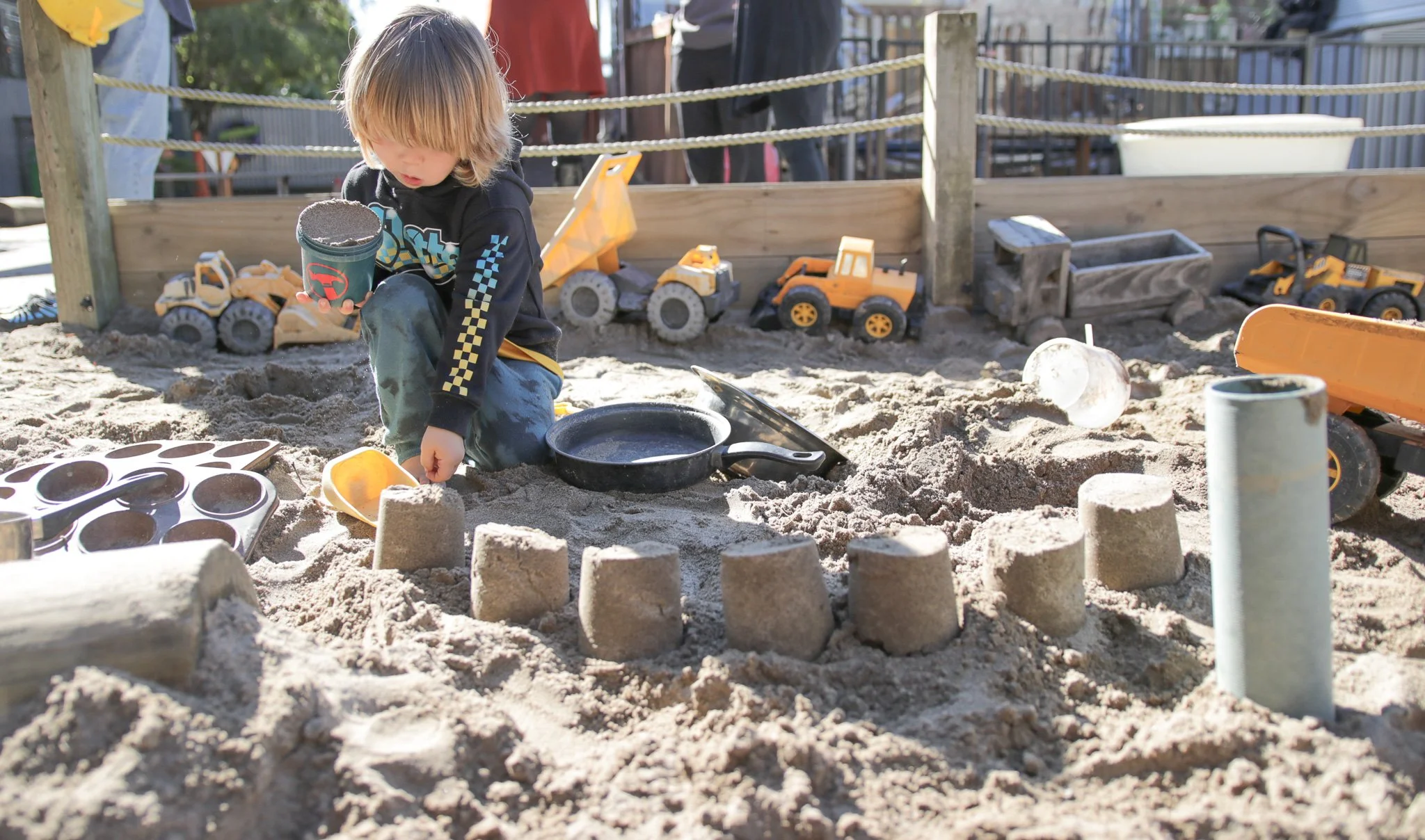 A young child with blonde hair playing in a sandy sandbox at a playground, surrounded by toy construction vehicles and sand molds, with a wooden fence and some people in the background, under sunlight.