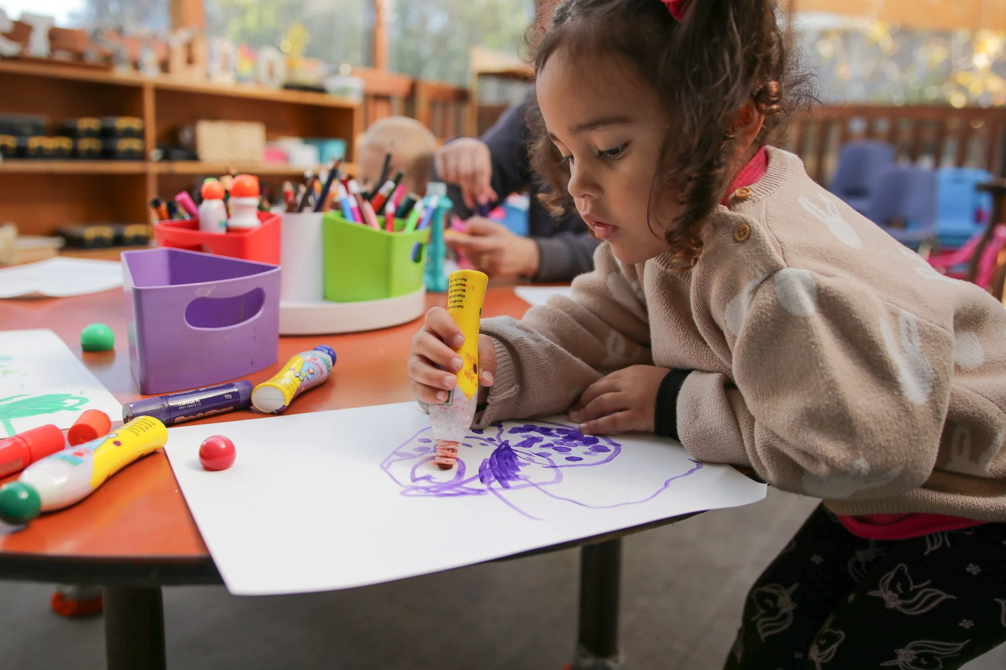 A young girl with curly hair drawing with purple marker on a white sheet of paper at a craft table filled with colorful art supplies in a bright room.