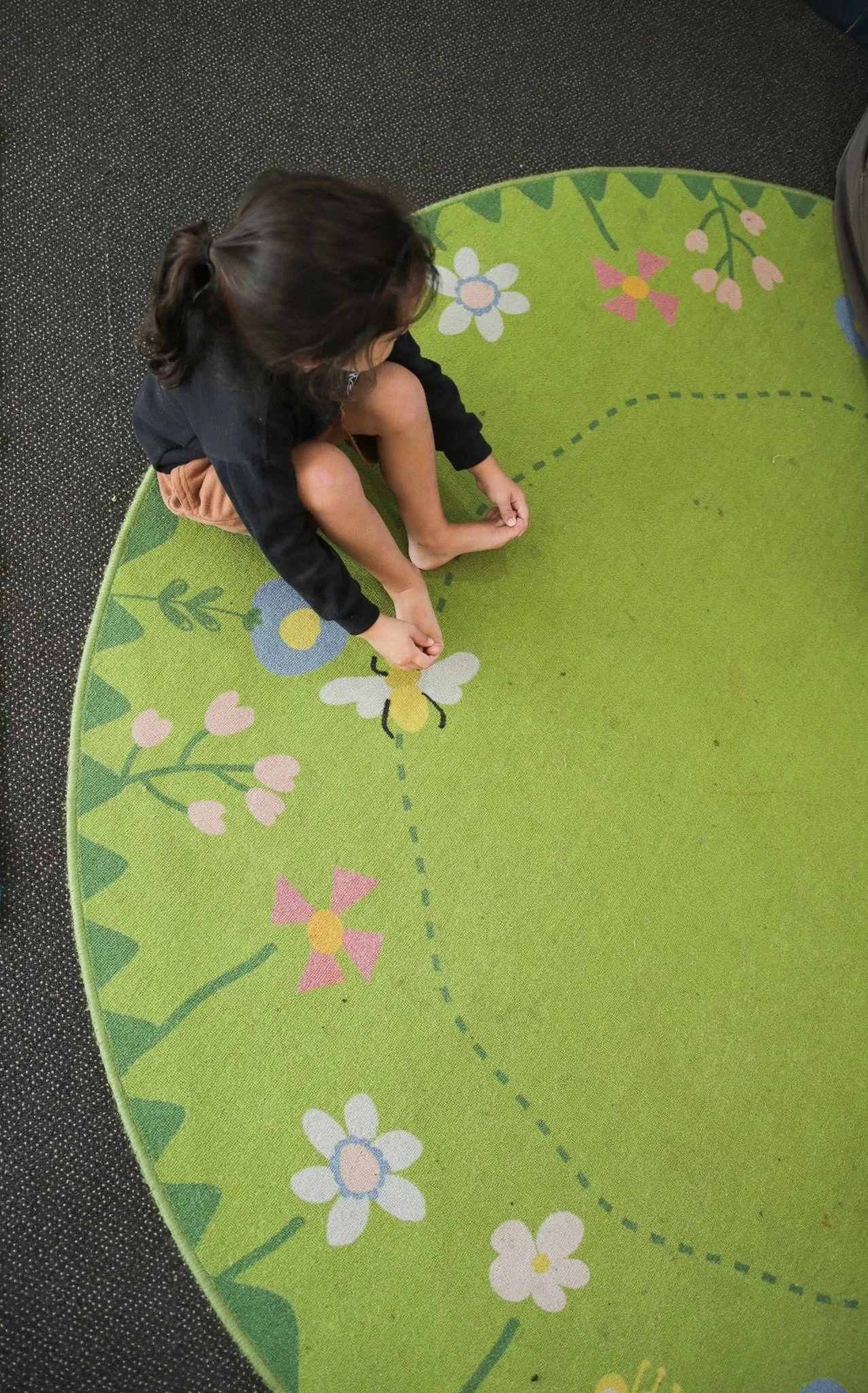 A young girl with dark hair tied back, sitting on a green circular rug with colorful flowers and butterflies, on a dark speckled floor.