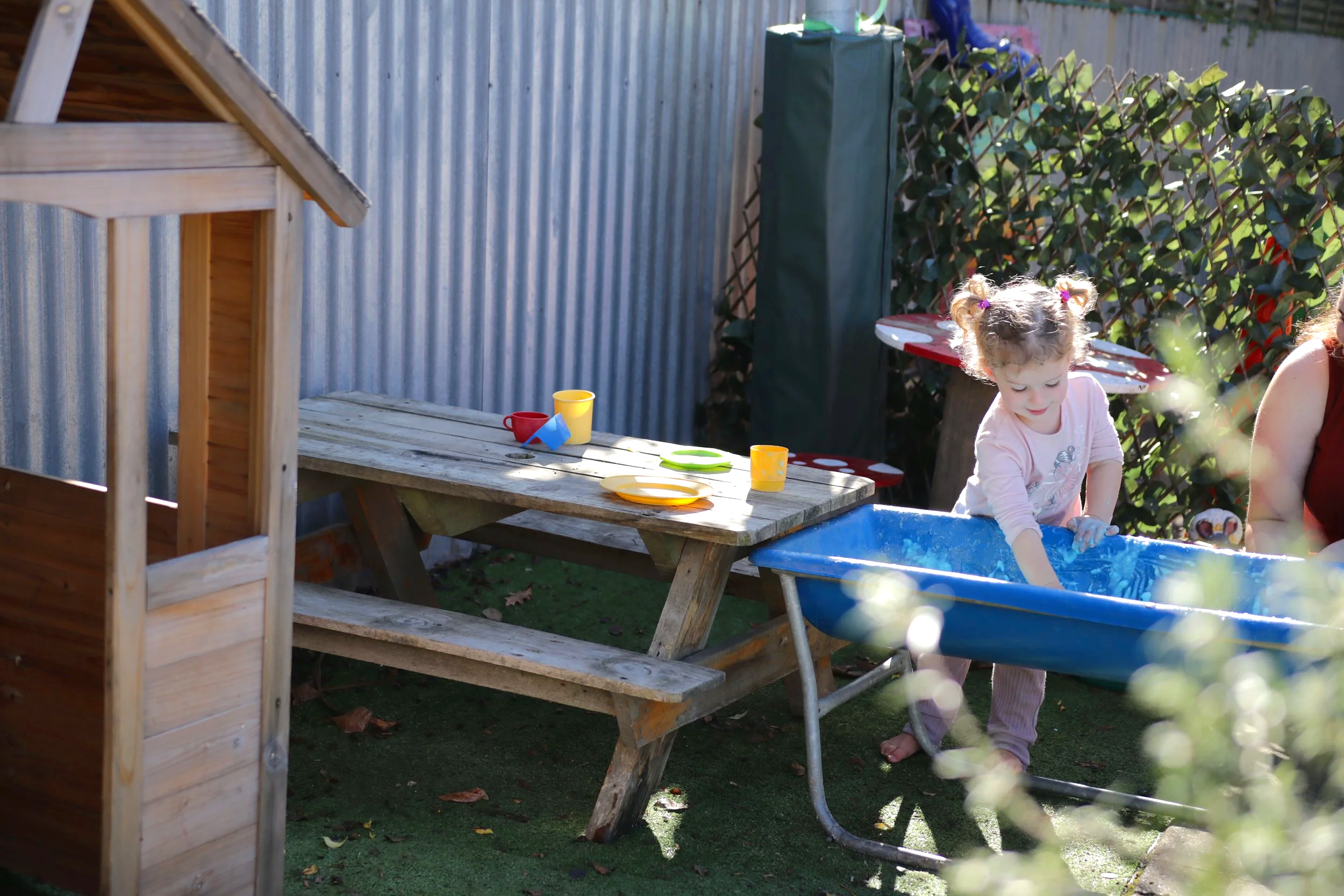 A young girl playing in a blue plastic kiddie pool in a backyard. There are colorful plastic cups and plates on a wooden picnic table nearby. The scene is sunny with greenery and a metal fence in the background.