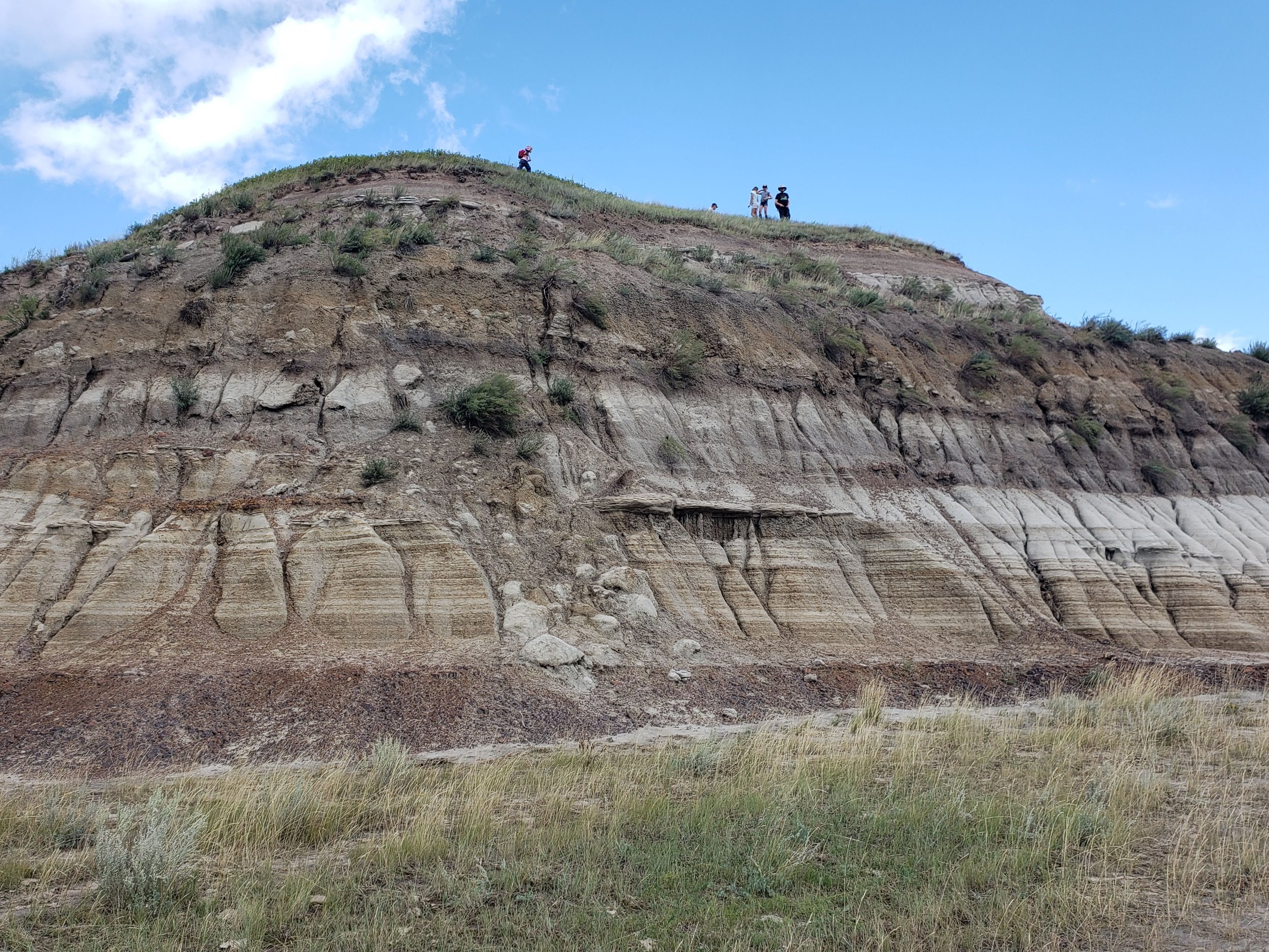 People walking along the top of a layered, rocky cliff with eroded patterns and sparse vegetation, under a partly cloudy sky.