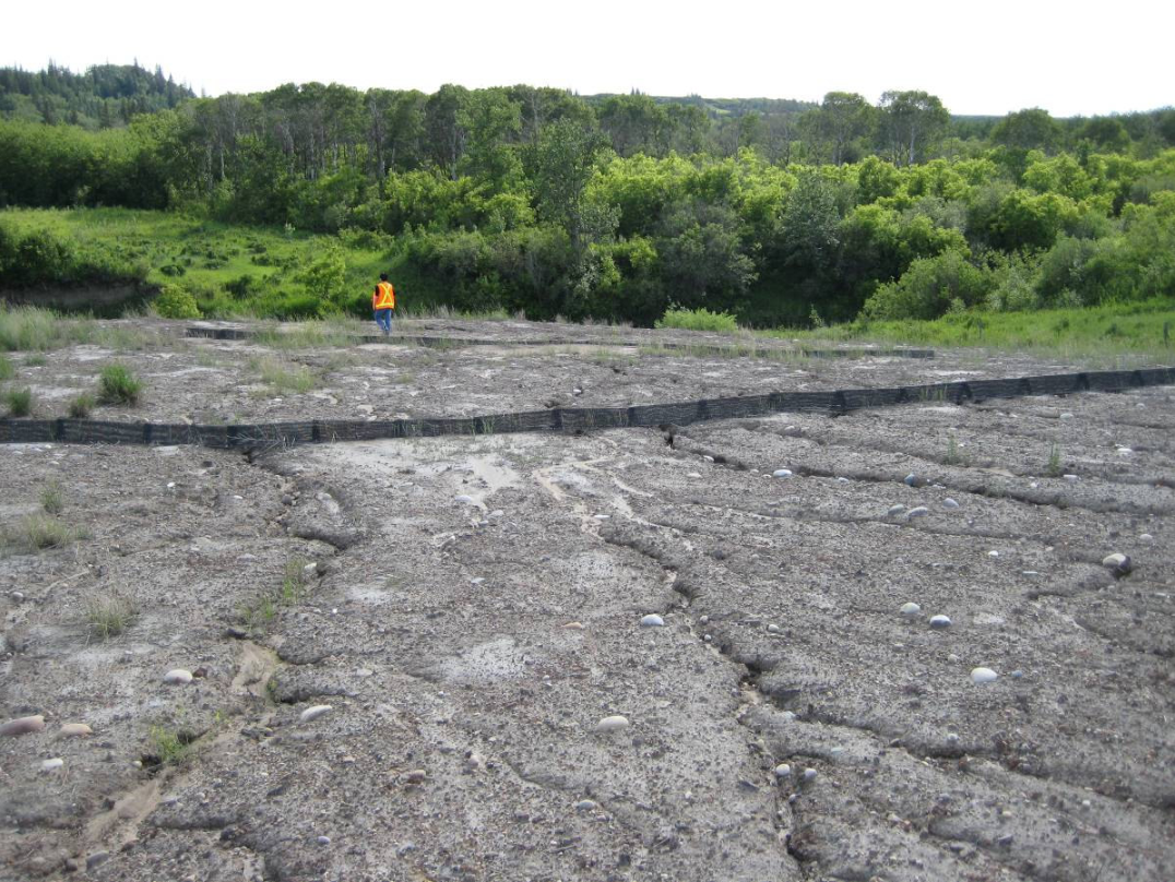 A person in a safety vest inspecting a landslide due to erosion, showing cracked and uneven ground with soil and small rocks, with a lush green forest in the background.