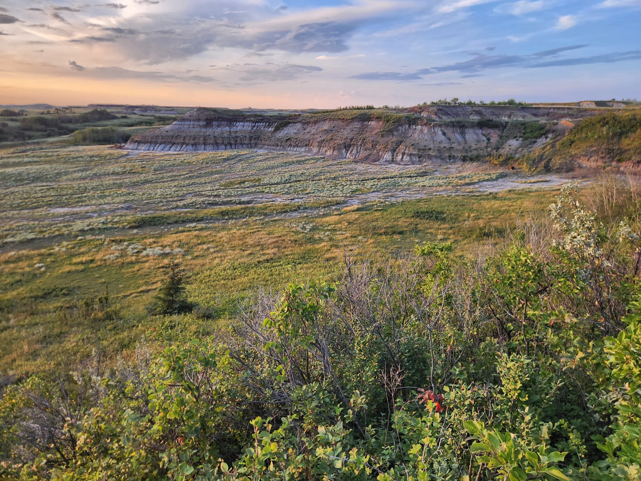 A landscape with a gently rolling terrain, showing a hill with layered rock formations under a partly cloudy sky at sunset or sunrise, which is under threat of destruction from sodium bentonite clay mining, industrial use on agricultural land.