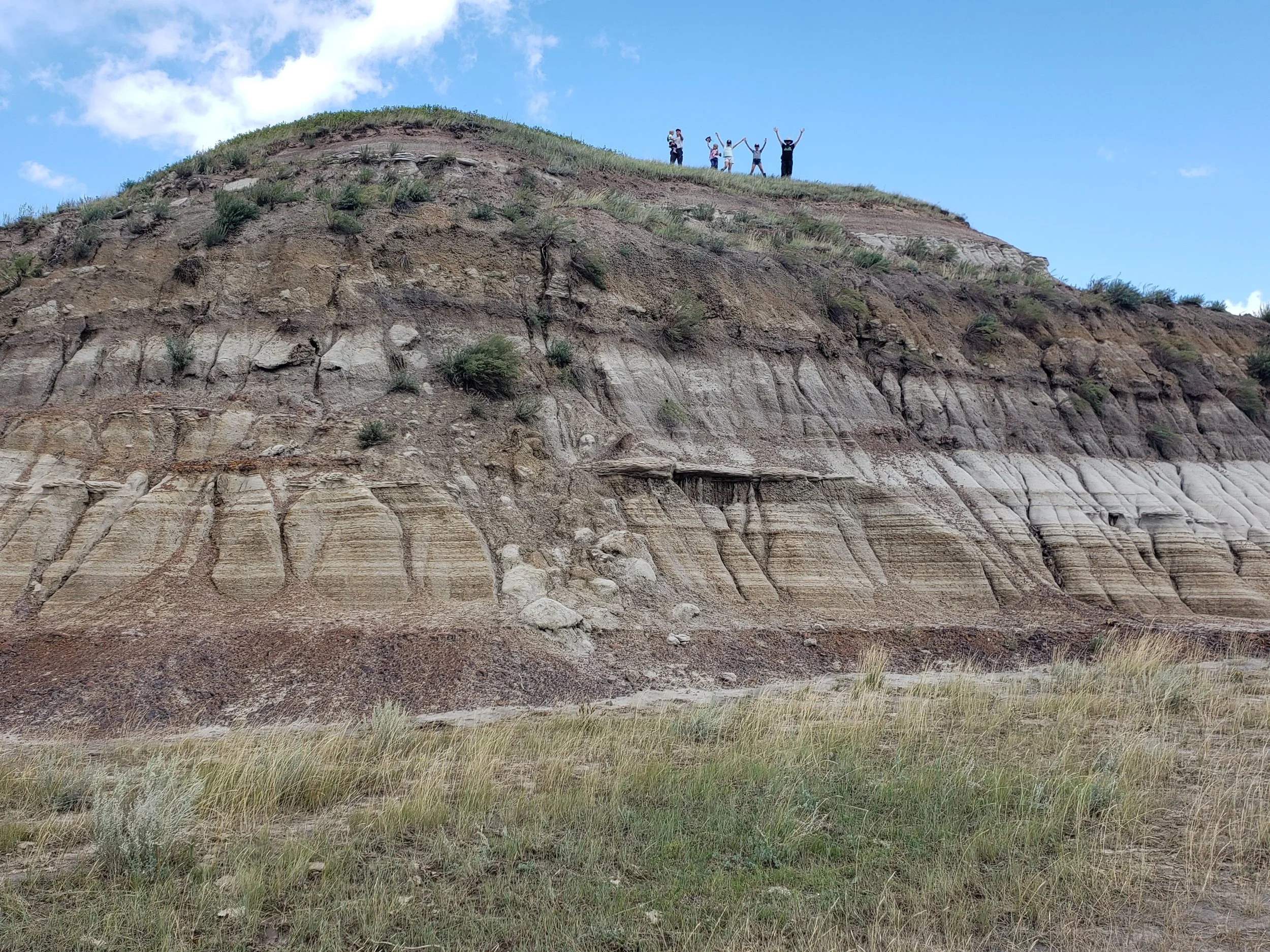 A group of six people standing on top of a tall badlands hill, with sodium bentonite layer, ironstone, and exposed bedrock, celebrating with their arms raised against a partly cloudy blue sky.