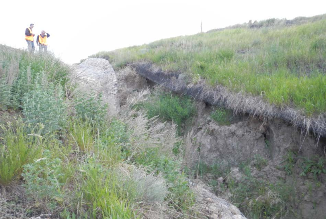 Three people in yellow vests examining a large ground collapse due to erosion on a grassy hillside.