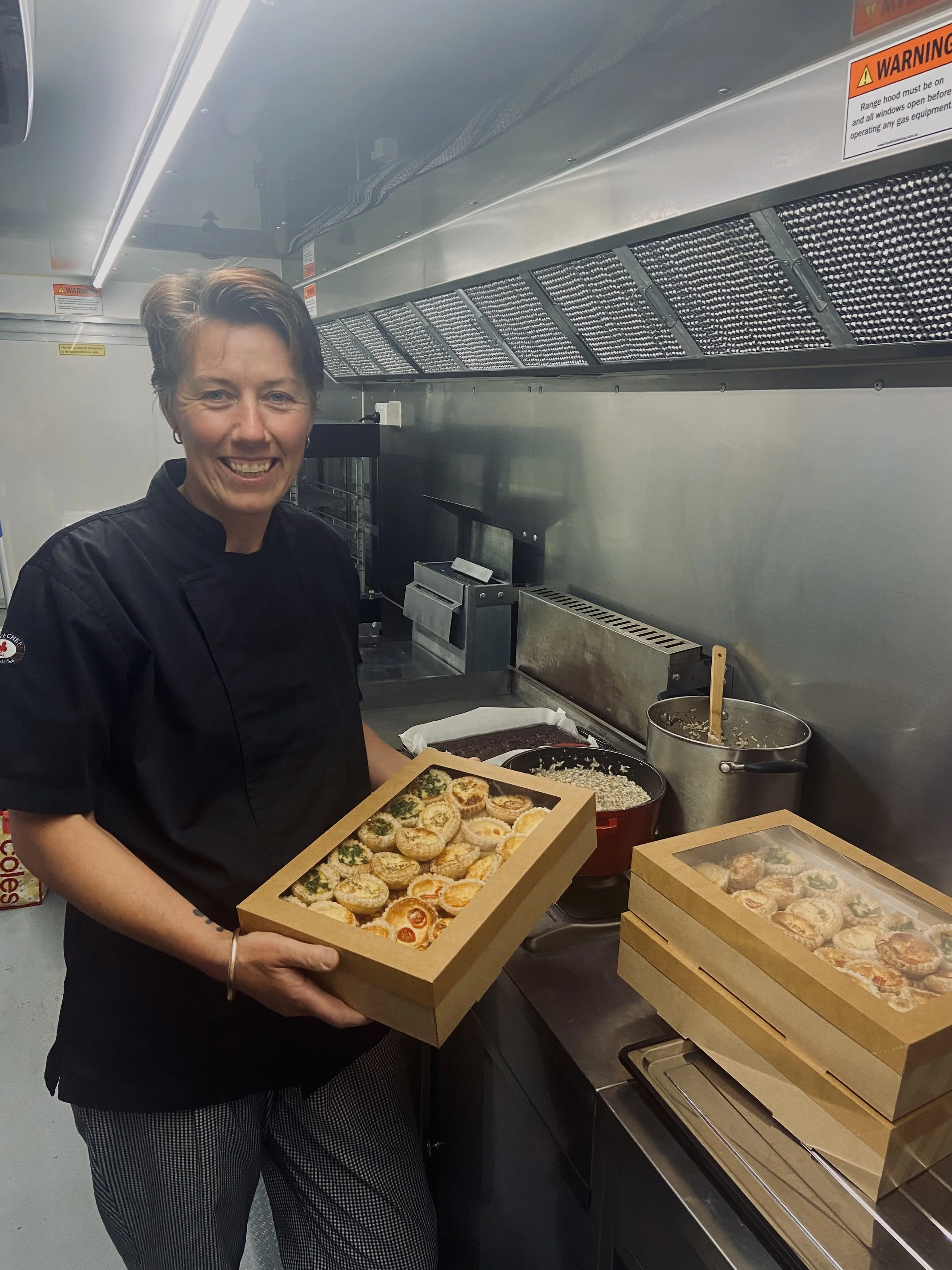 A smiling chef holding a tray of mini savory tarts in a commercial kitchen.