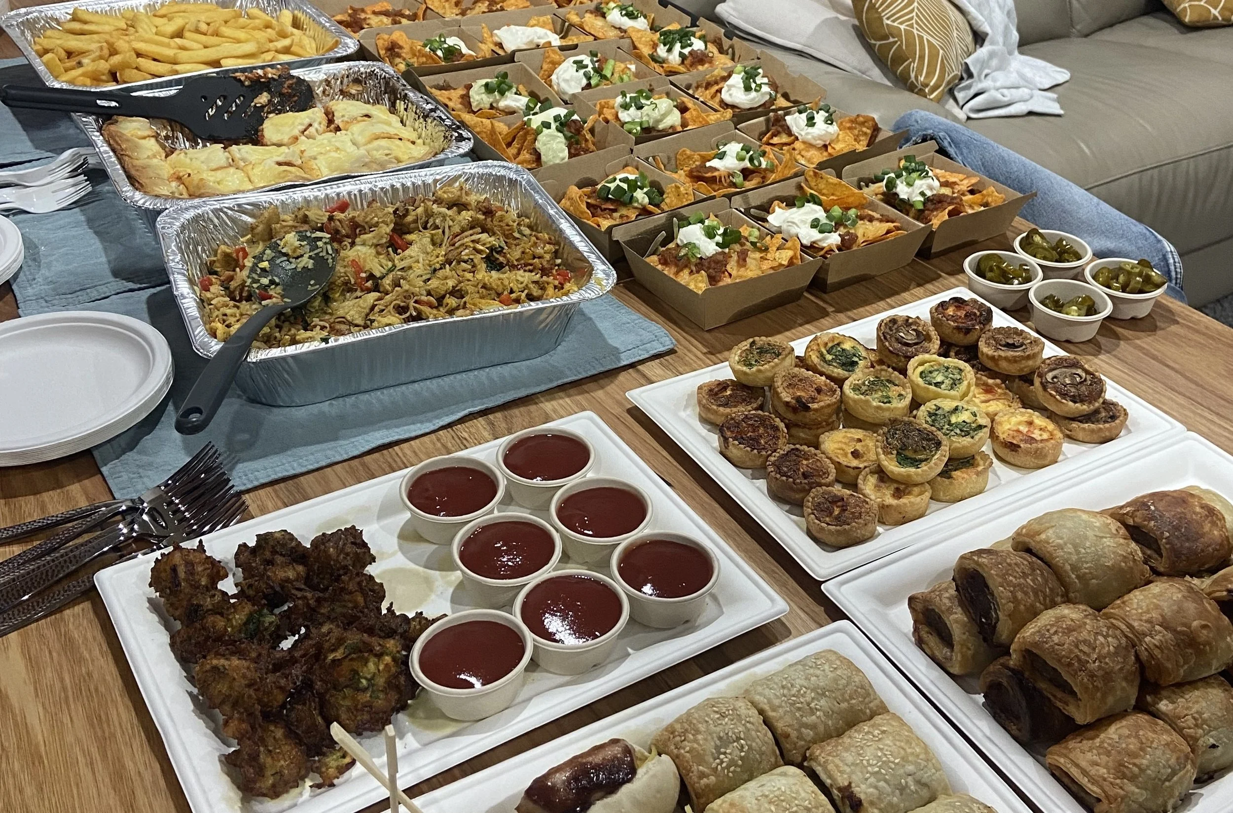 A table filled with various party foods including French fries, baked chicken wings, a casserole dish, small taco salads with sour cream and green onions, mini quiches, muffins, and several small bowls of red sauce and jalapeños, set in a casual living room.