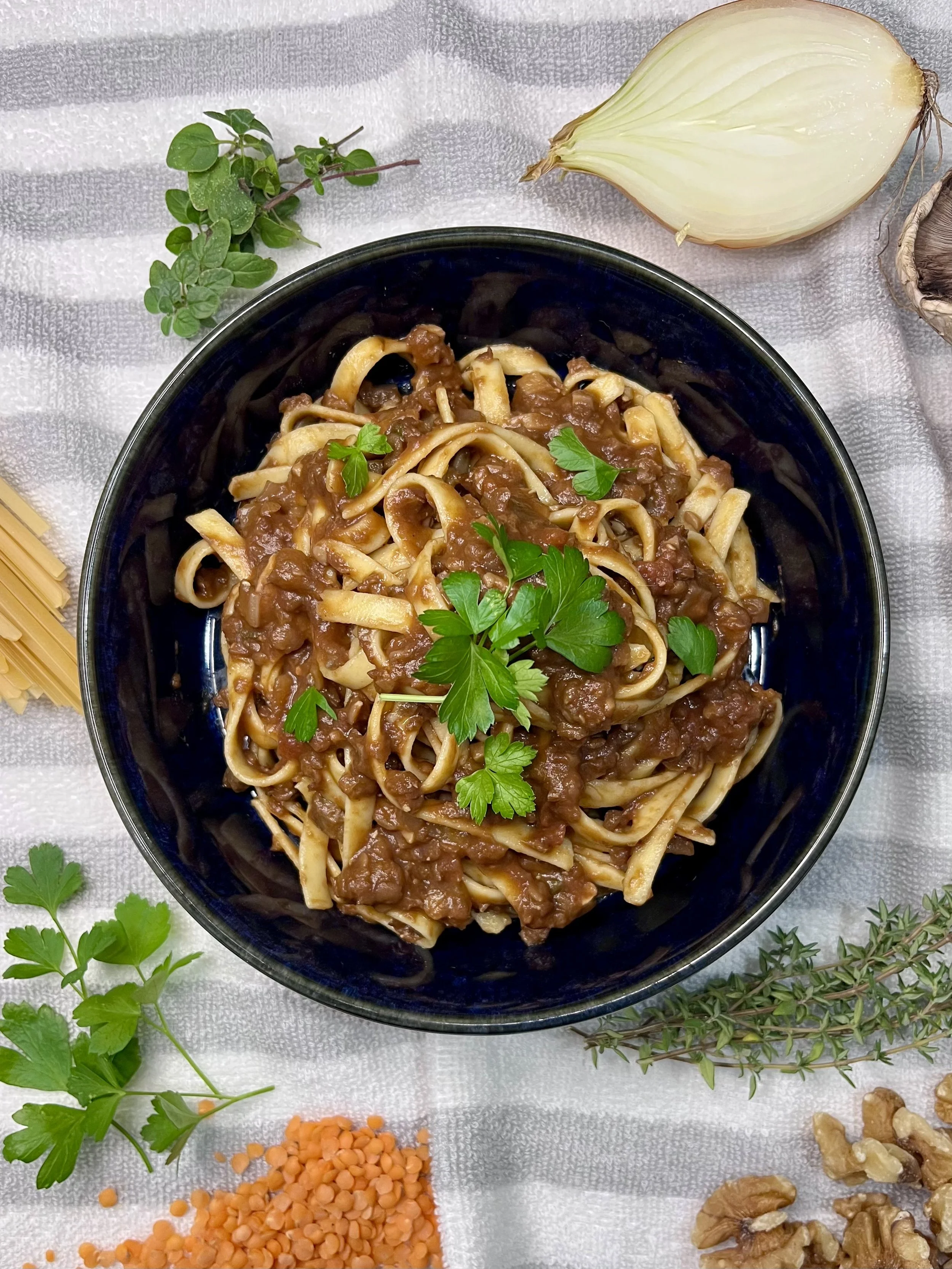 A bowl of cooked fettuccine pasta with tomato-based vegan bolognaise sauce garnished with fresh parsley, surrounded by fresh herbs, onion, walnuts, and lentils on a white textured cloth.