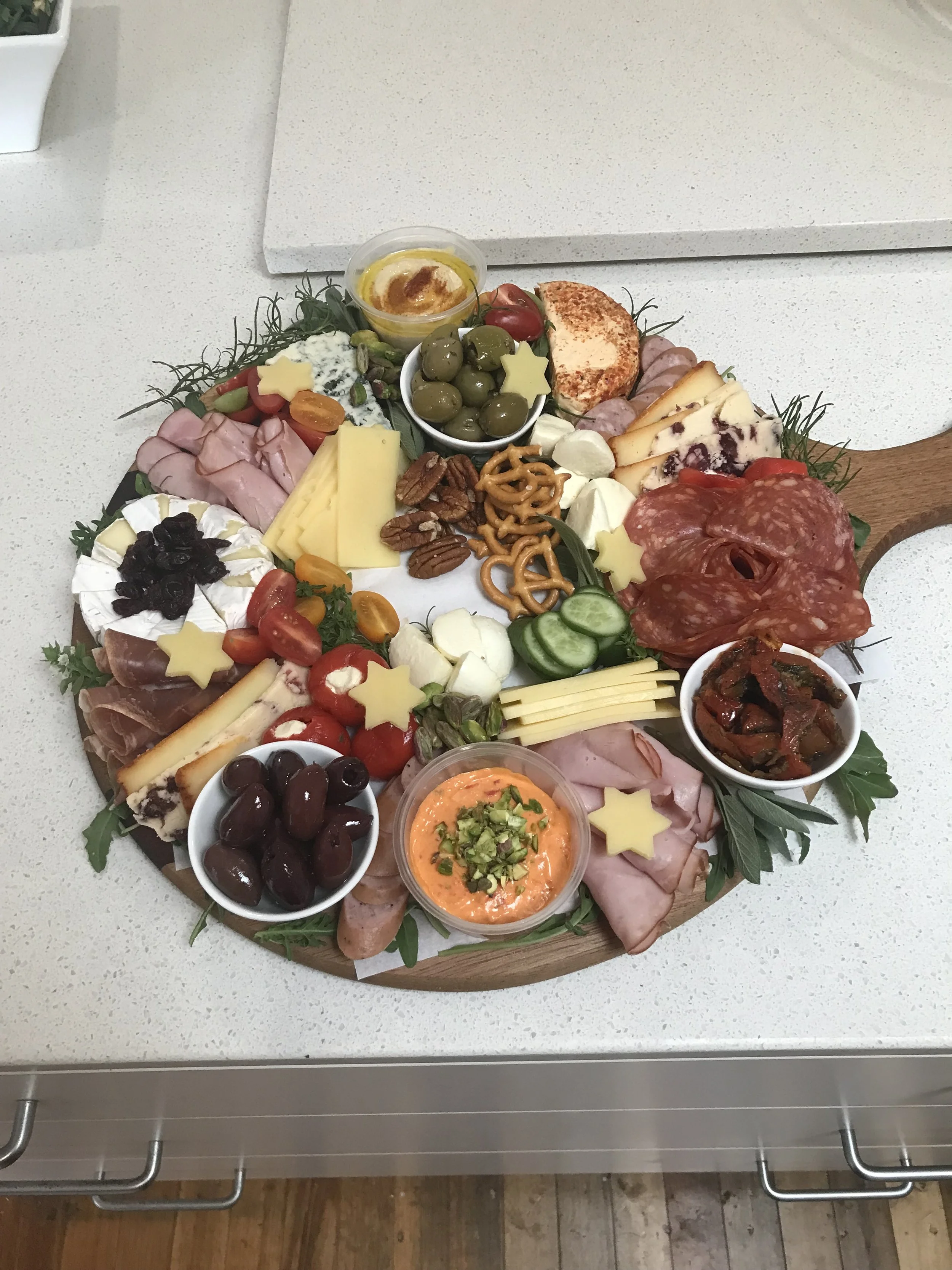 A cheese and charcuterie board with assorted cheeses, meats, olives, cherry tomatoes, nuts, dips, and garnishes, placed on a round wooden platter on a white table.
