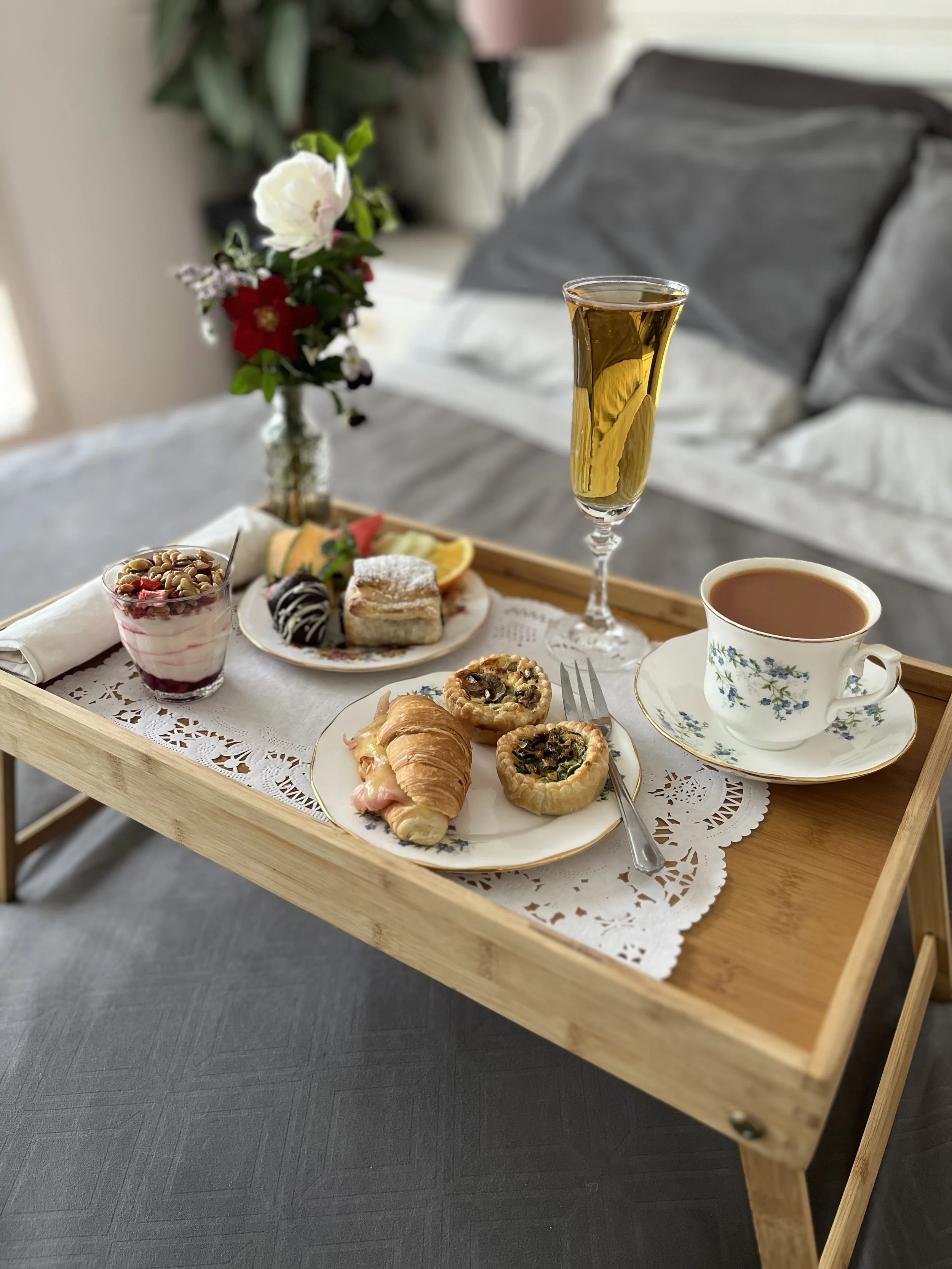 A wooden tray with a lace doily holding a breakfast setup, including a glass of champagne, a cup of tea, a small yoghurt and pastries, with a floral arrangement in the background.
