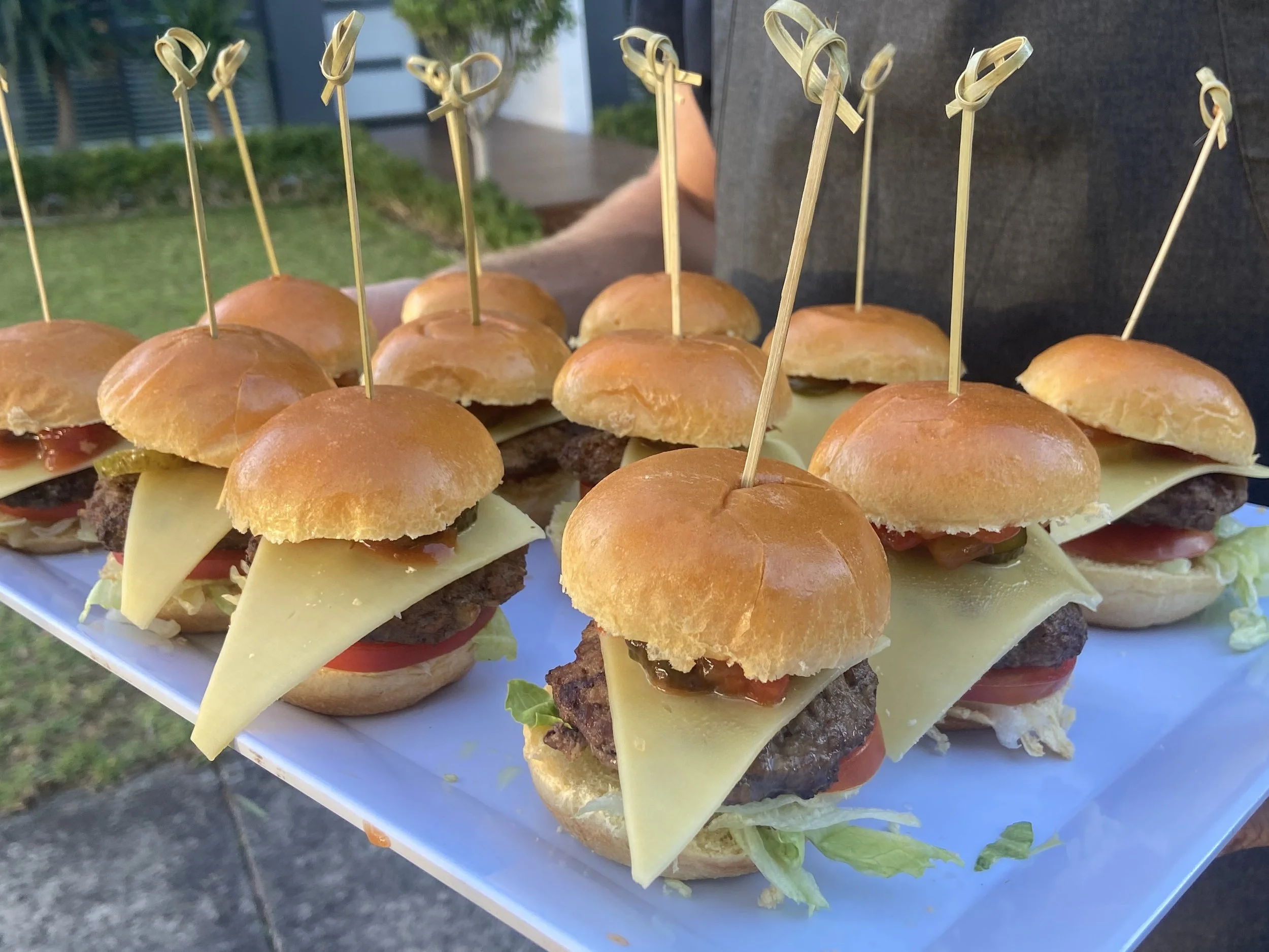 A tray of miniature hamburgers topped with cheese, lettuce, tomato, and onion, each held together with a skewer, outdoor setting with blurred greenery and buildings in the background.