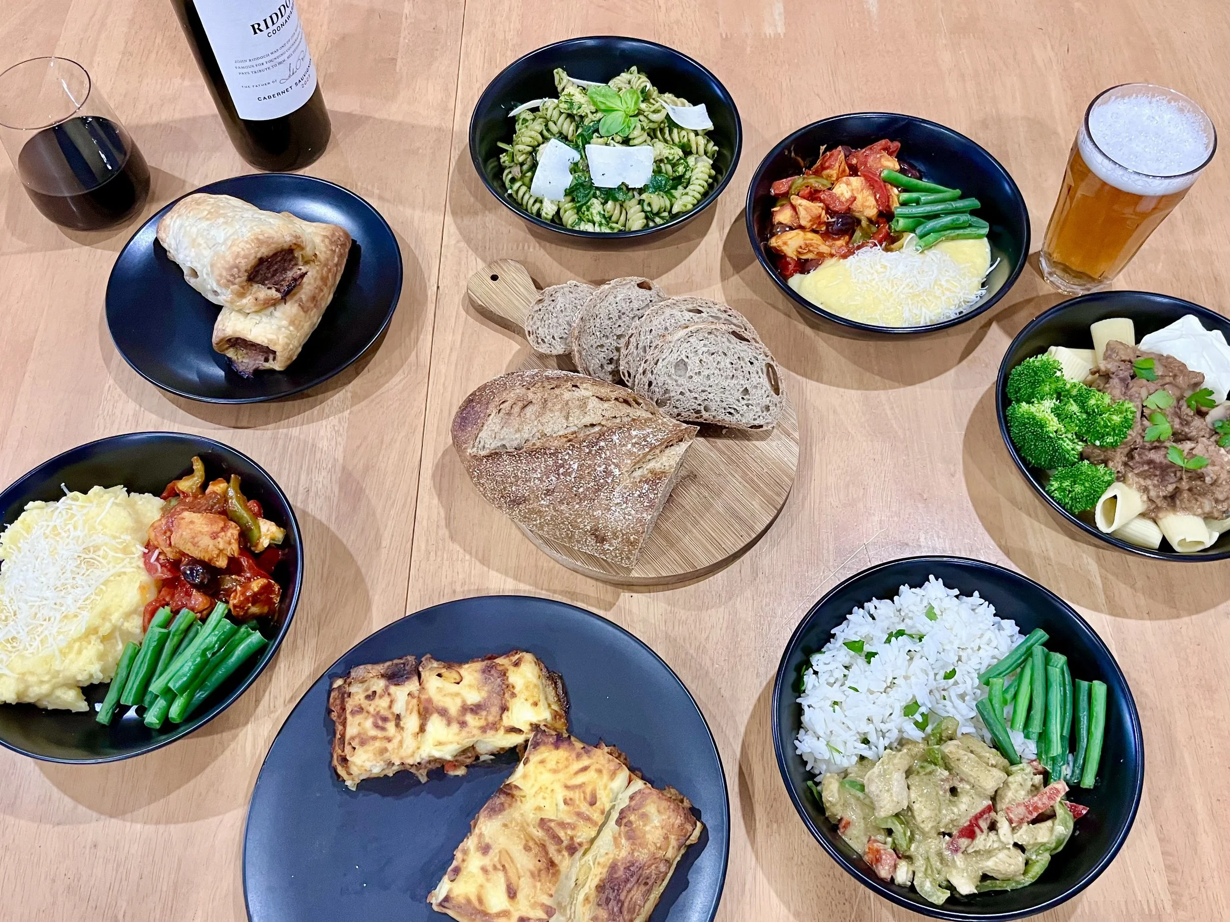 A table filled with various dishes including pasta, bread, baked casseroles, rice, and vegetable dishes, along with a bottle of red wine, a glass of red wine, and a glass of beer.