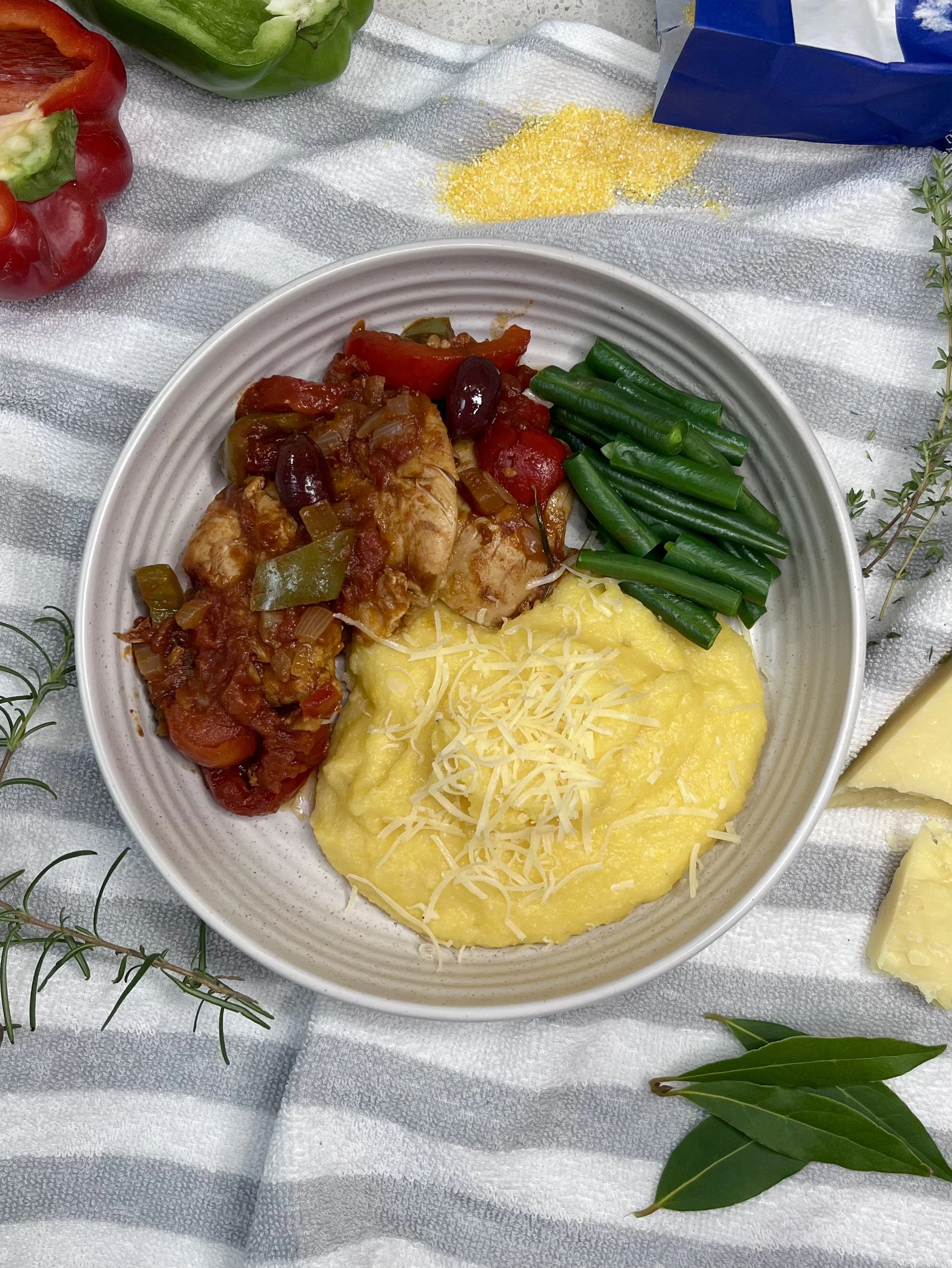 Plate with creamy polenta topped with shredded cheese, green beans, and a chicken cacciatore and vegetables, on a cloth with herbs and pepper.