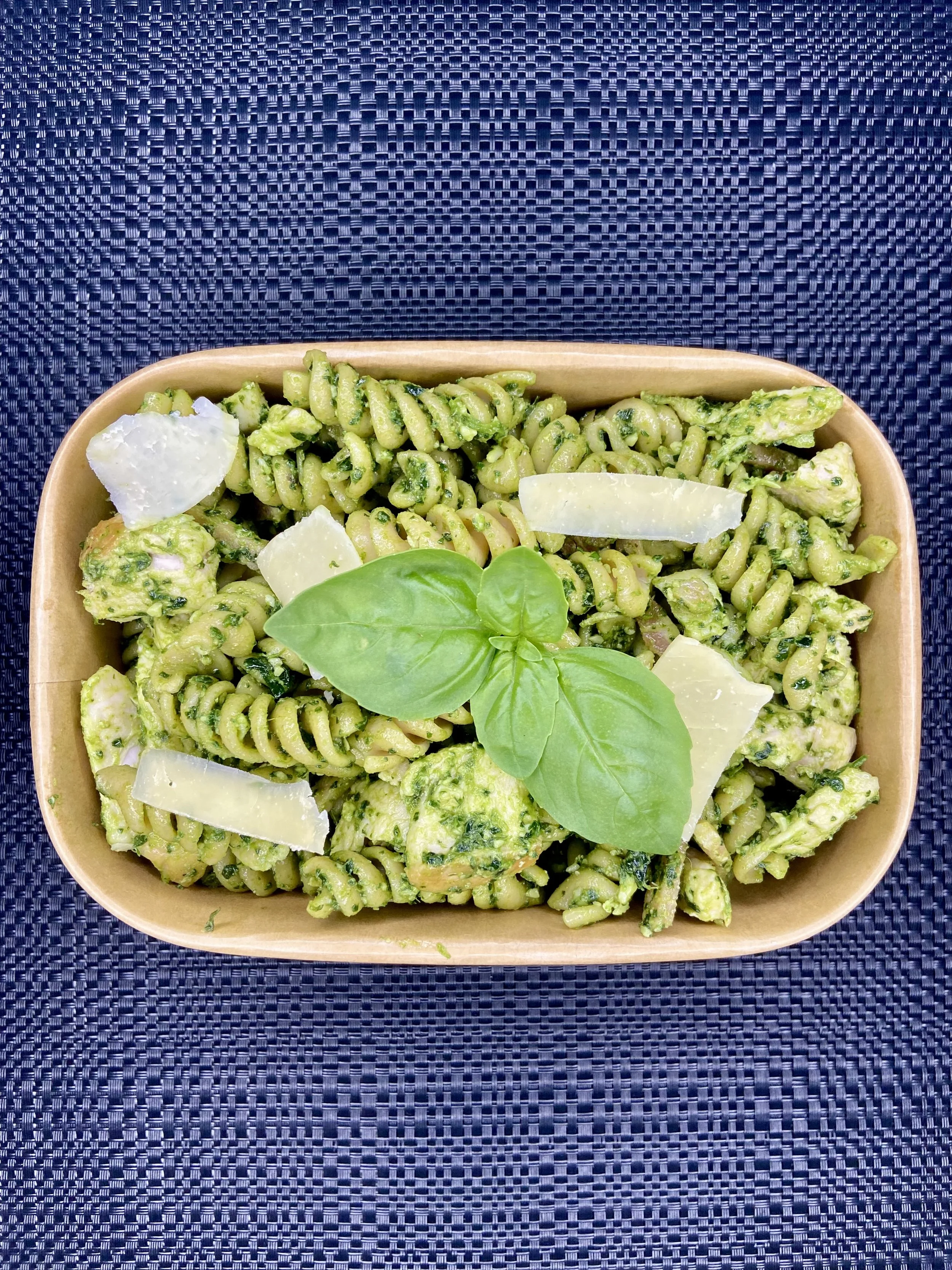 Fresh green pesto pasta in a brown takeaway container garnished with basil leaves and parmesan cheese slices, placed on a dark woven placemat.
