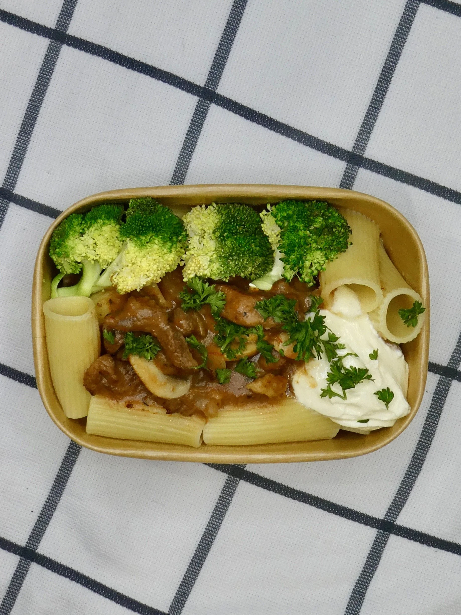 A rectangular takeaway container filled with pasta, broccoli florets, and beef stroganoff, garnished with parsley, on a white tablecloth with black grid pattern.