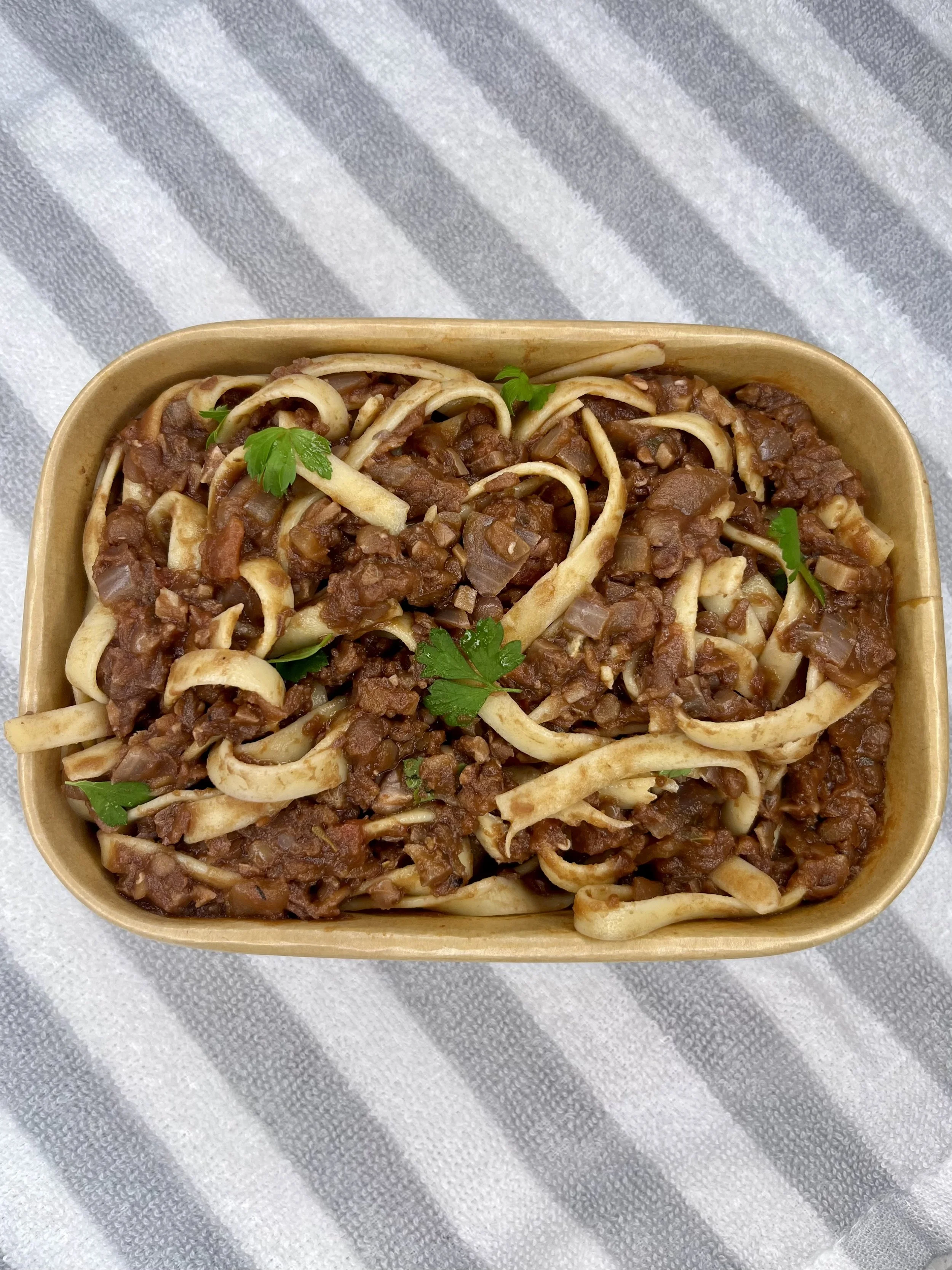 A rectangular takeout container filled with spaghetti topped with a meaty tomato sauce and garnished with fresh parsley, placed on a white and grey striped cloth.