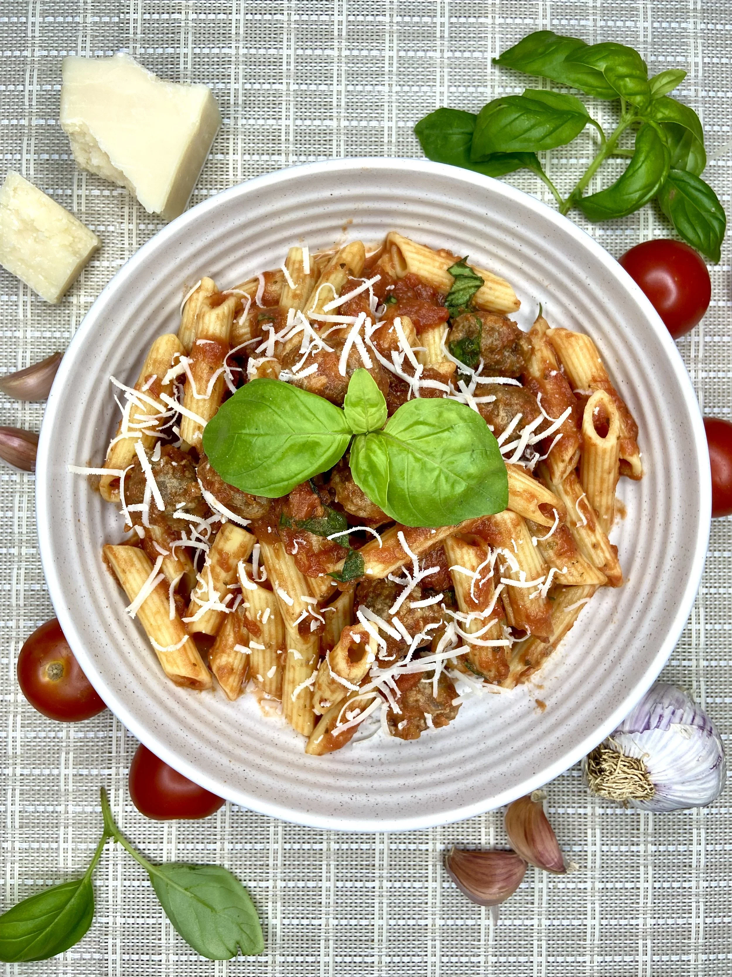 A plate of cooked pasta with tomato sauce topped with shredded cheese and a basil leaf, surrounded by cherry tomatoes, garlic cloves, and basil leaves on a woven placemat.