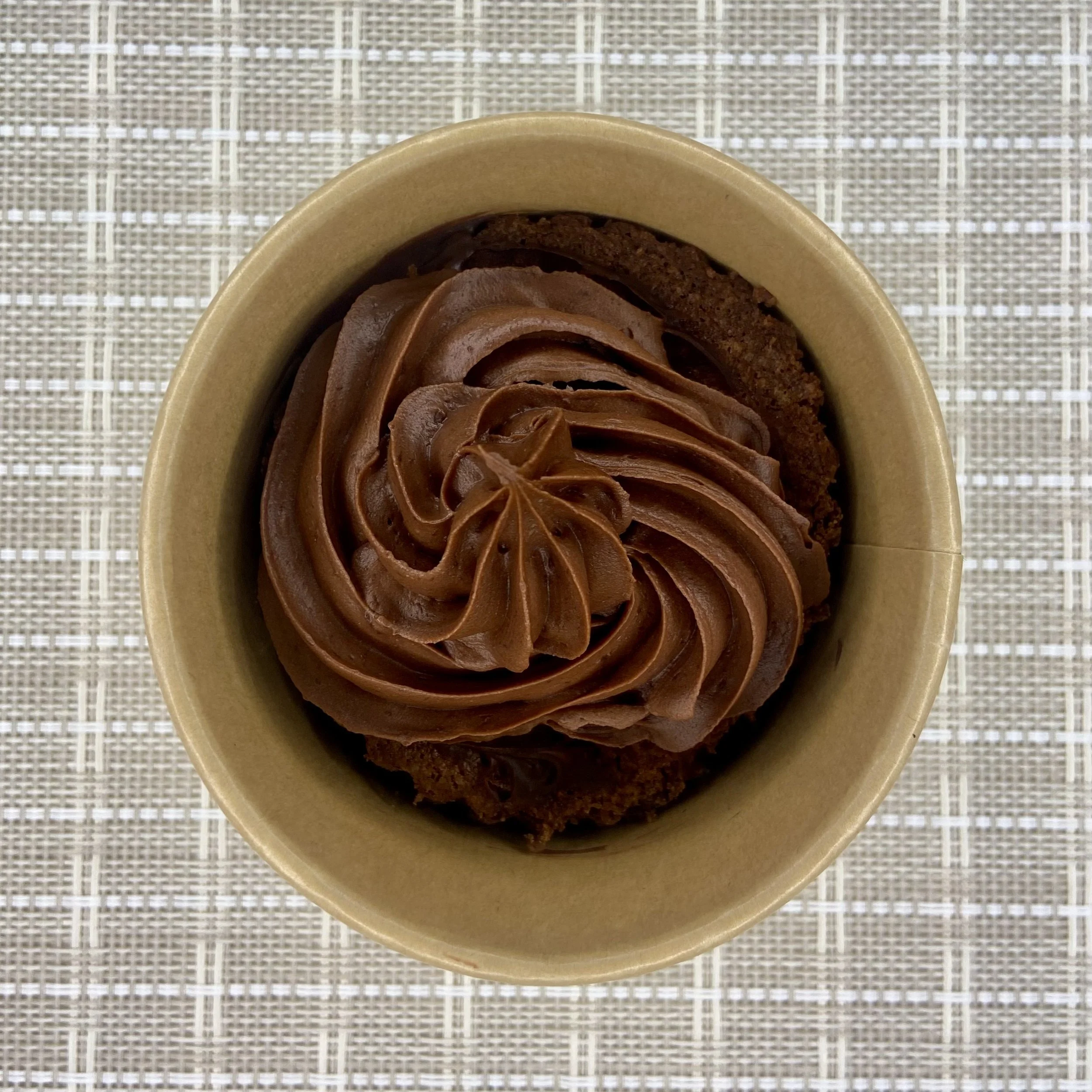 Close-up of a dessert featuring chocolate whipped topping in a small, beige paper cup on a woven textured surface.