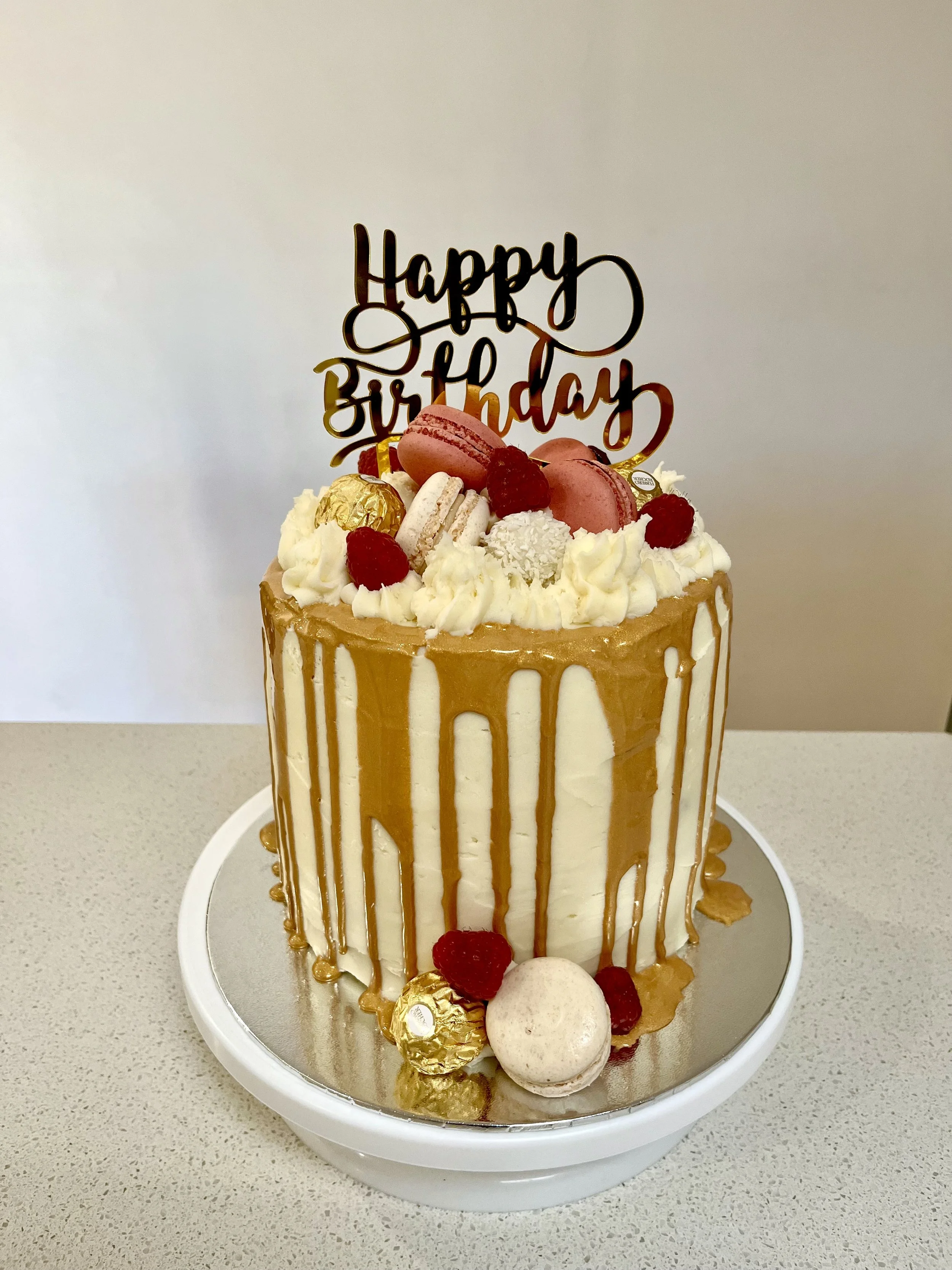 A tall birthday cake with white icing and caramel drizzle, topped with macarons, raspberries, and chocolates, with a 'Happy Birthday' cake topper, sitting on a silver cake board on a beige countertop.