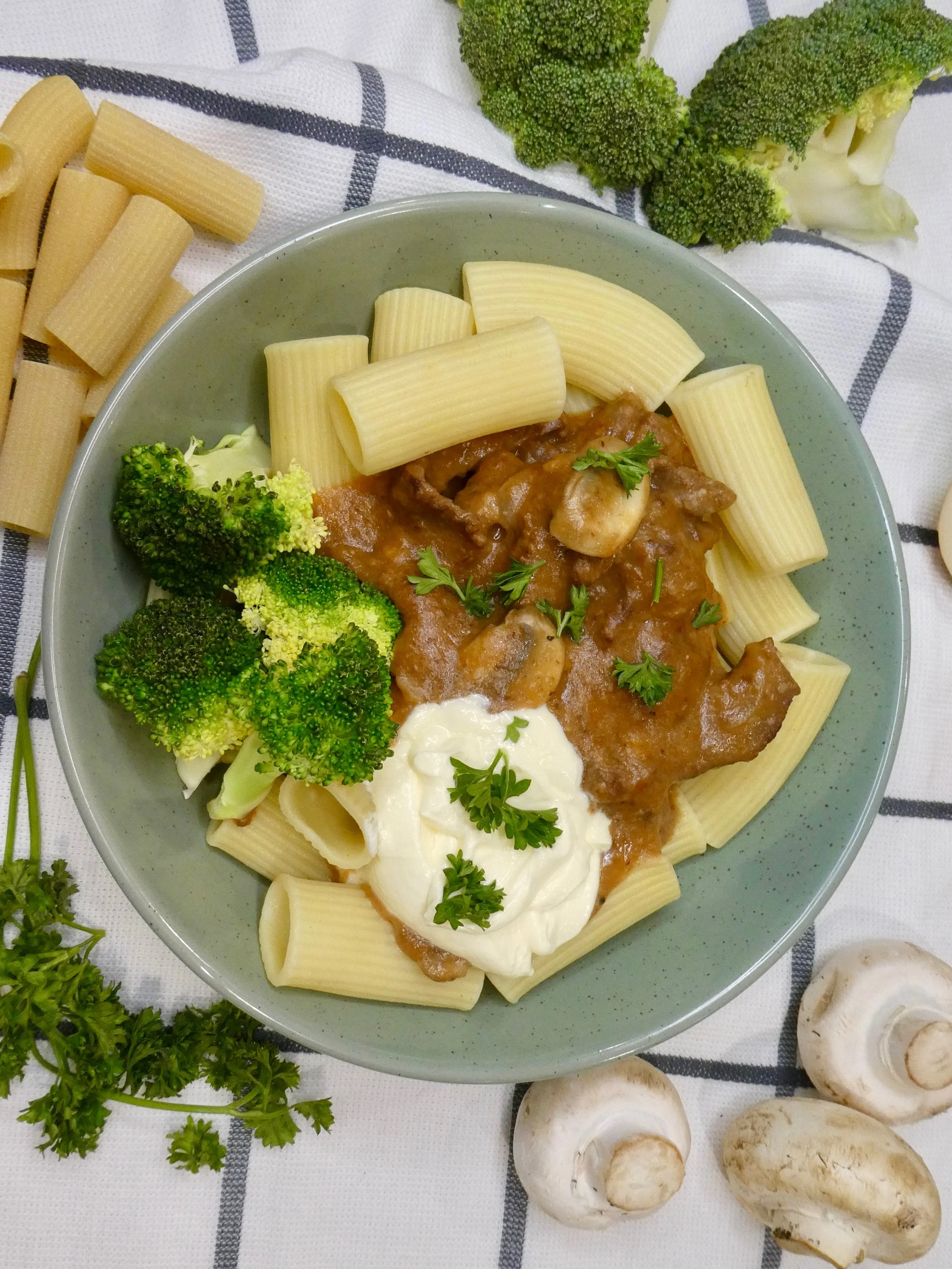 A bowl of baked pasta with broccoli, mushrooms, and a meat sauce, garnished with parsley, surrounded by fresh raw vegetables and mushrooms on a checkered cloth.