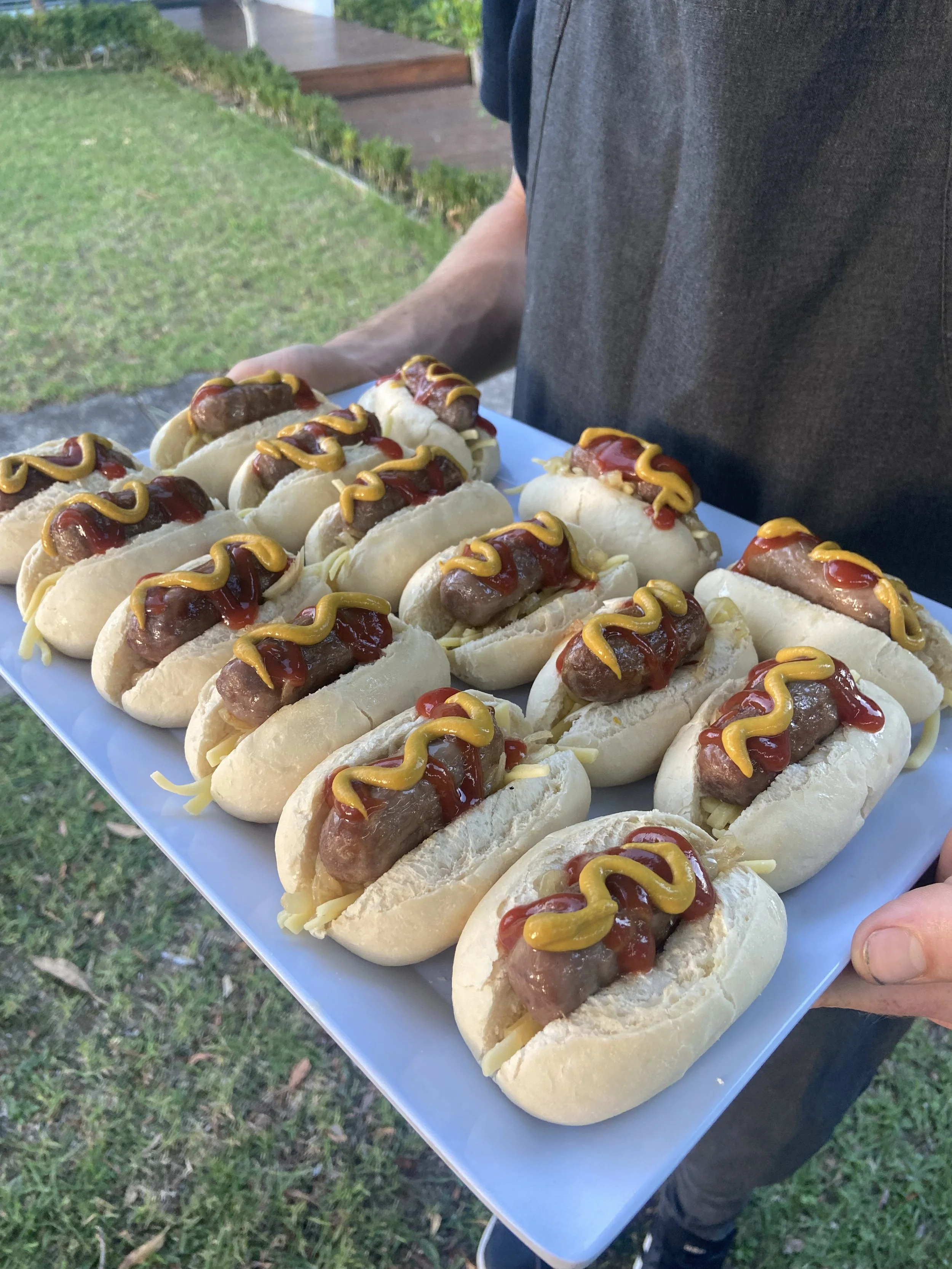 A person holding a white tray filled with hot dogs topped with ketchup and mustard outside on grass in a garden.