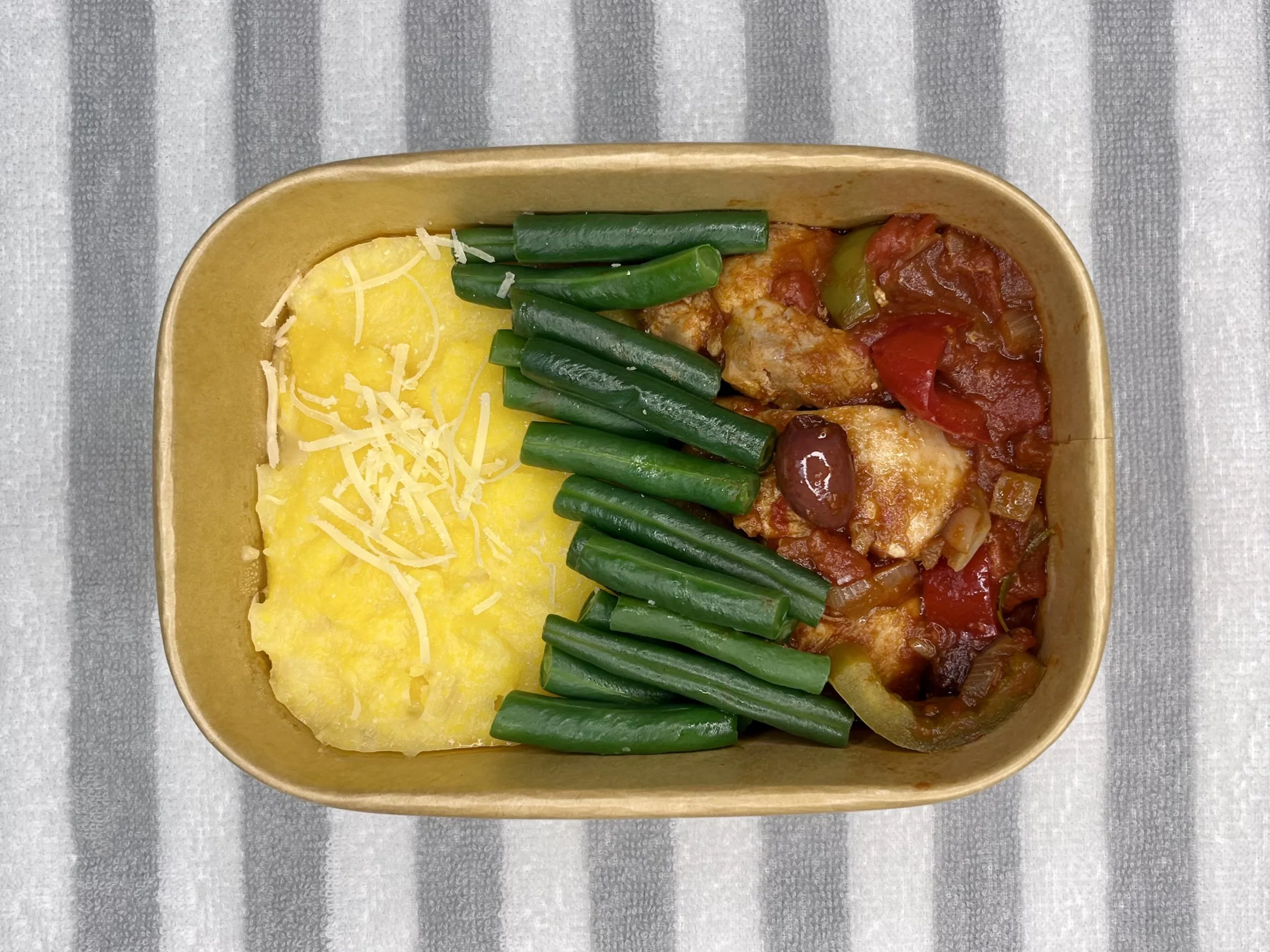 A rectangular takeaway carton containing creamy polenta topped with shredded cheese, cooked green beans, a piece of cooked chicken with tomato sauce, and sliced green peppers, placed on a gray and white striped cloth.