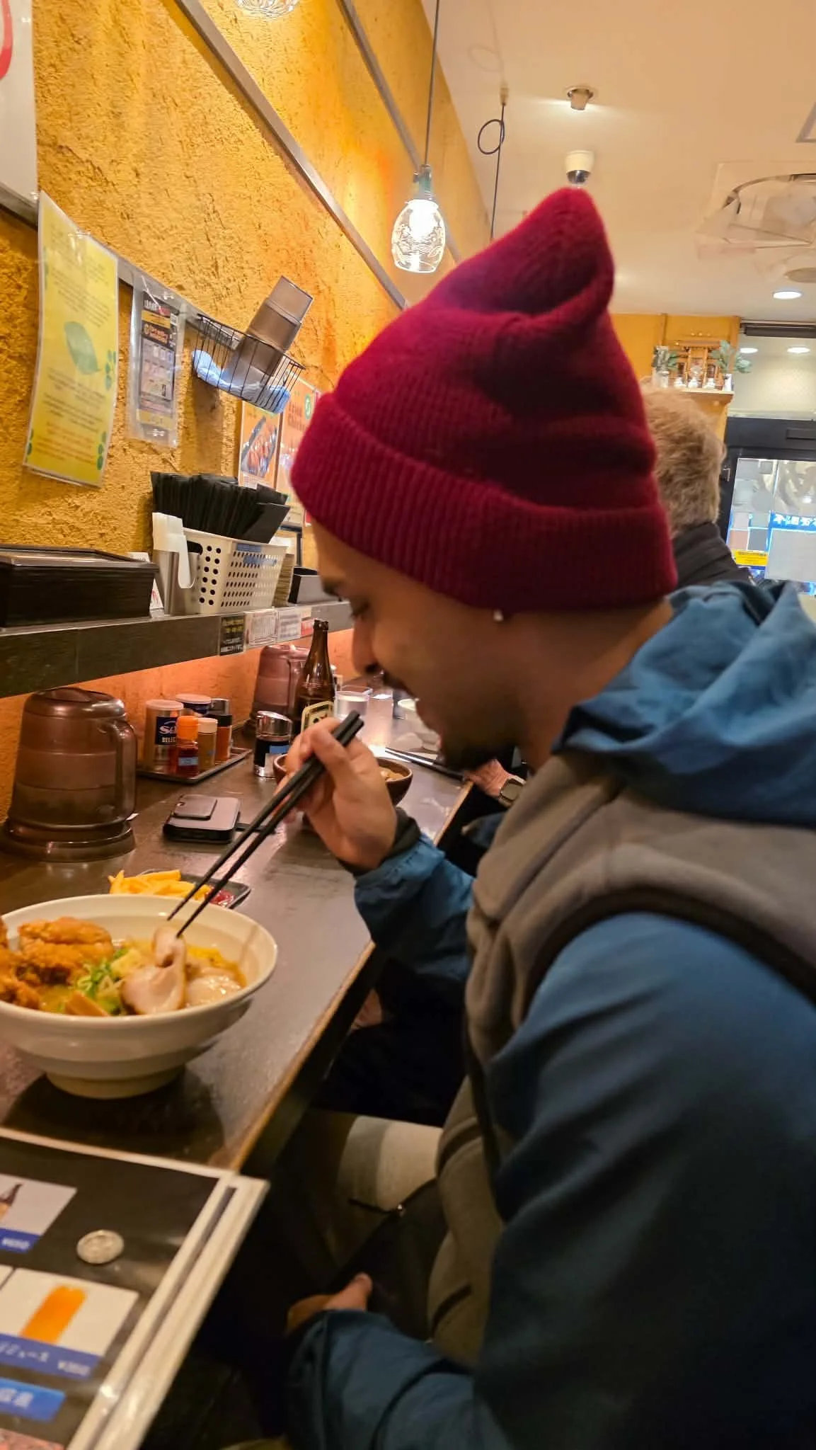 A person wearing a red beanie and blue jacket sitting at a restaurant counter, eating ramen with chopsticks, with condiments and a menu visible on the counter, and yellow textured walls in the background.