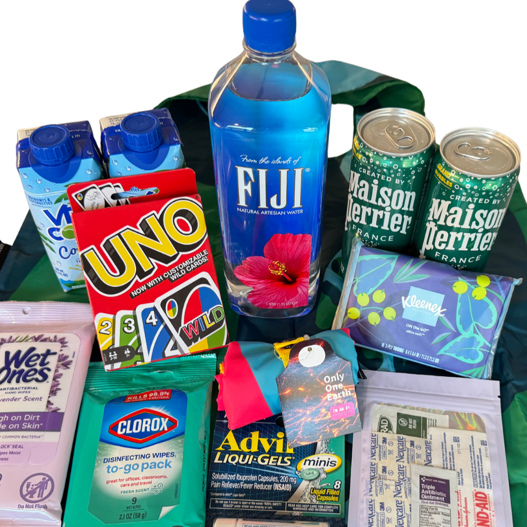 Assorted travel essentials including bottled water, other beverages, disinfecting wipes, pain relief gels, tissues, UNO cards, wipes, and snacks, all arranged in a reusable Hotel Arrival bag.
