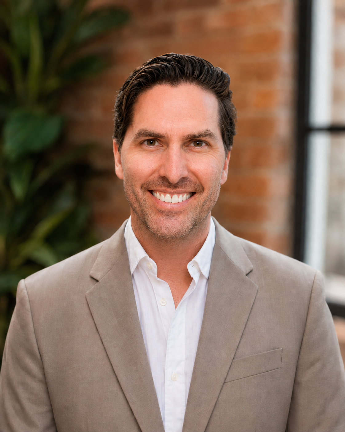 A smiling man in a beige suit and white shirt, with dark, styled hair, standing indoors with green plants and a brick wall in the background.