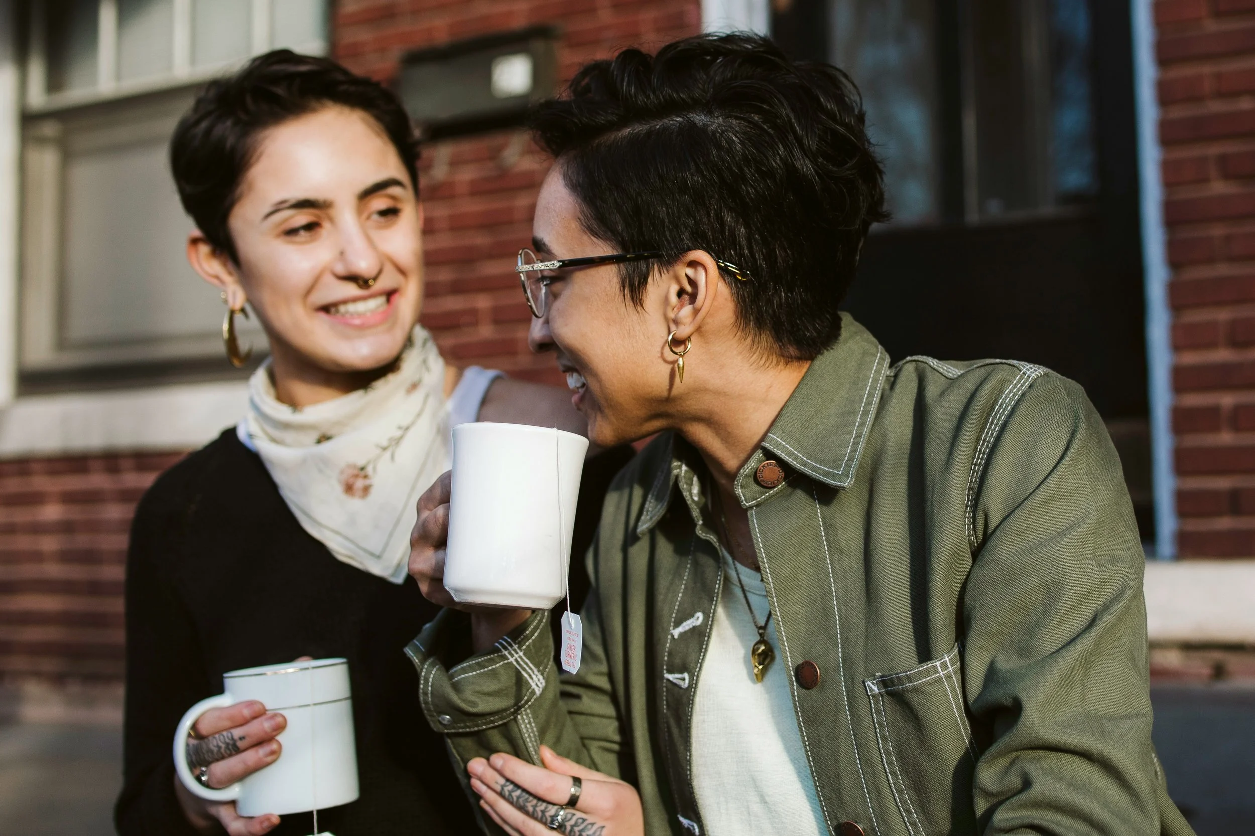 two adults of different races sit side by side looking at each other while holding mugs. Both have very short hair, dangling earrings and have matching tattoos on their middle fingers