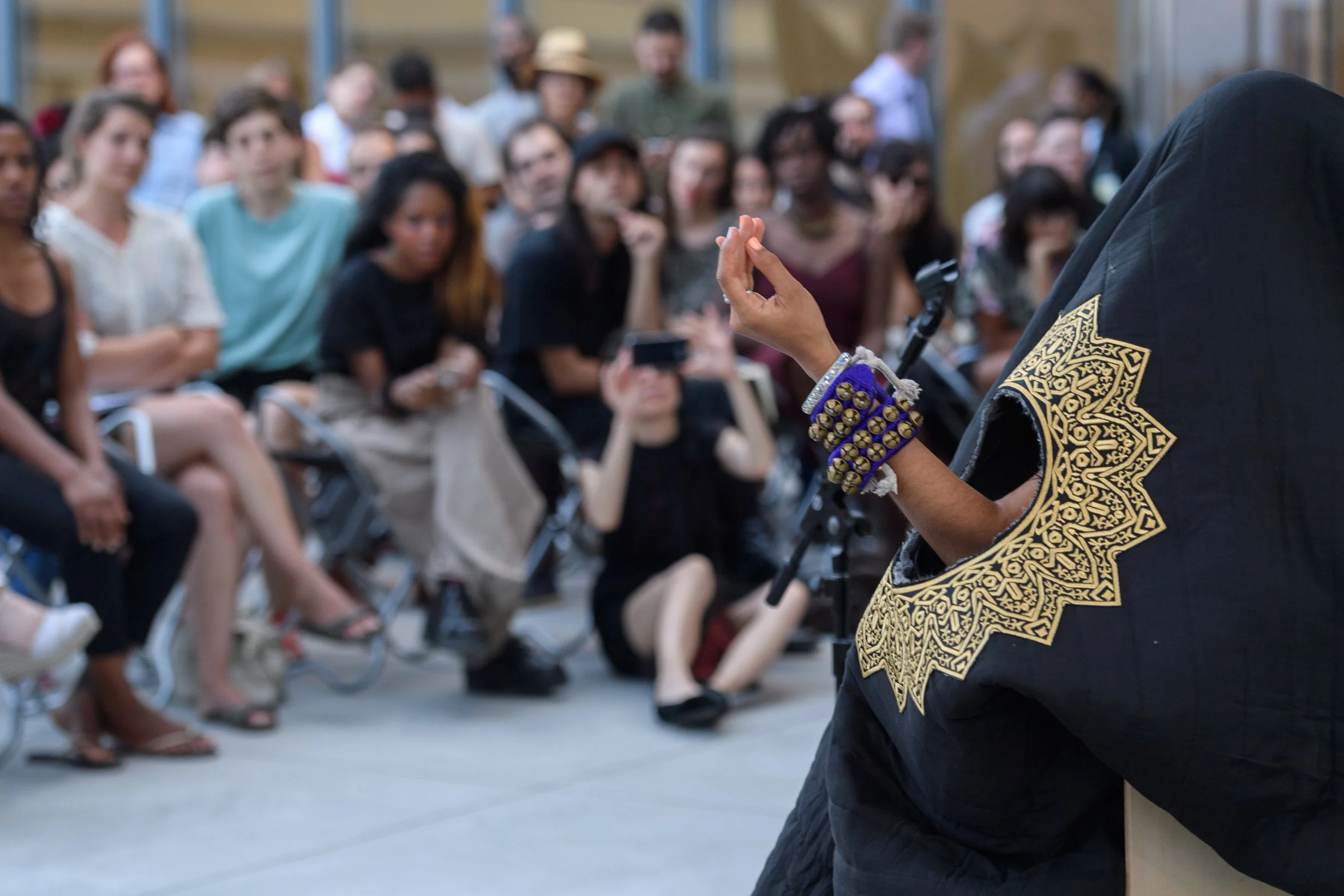 a brown skinned arm with bells on the wrist is held up through a hole in a black blanket.  There hole is embroidered with gold in the shape of a petals.
