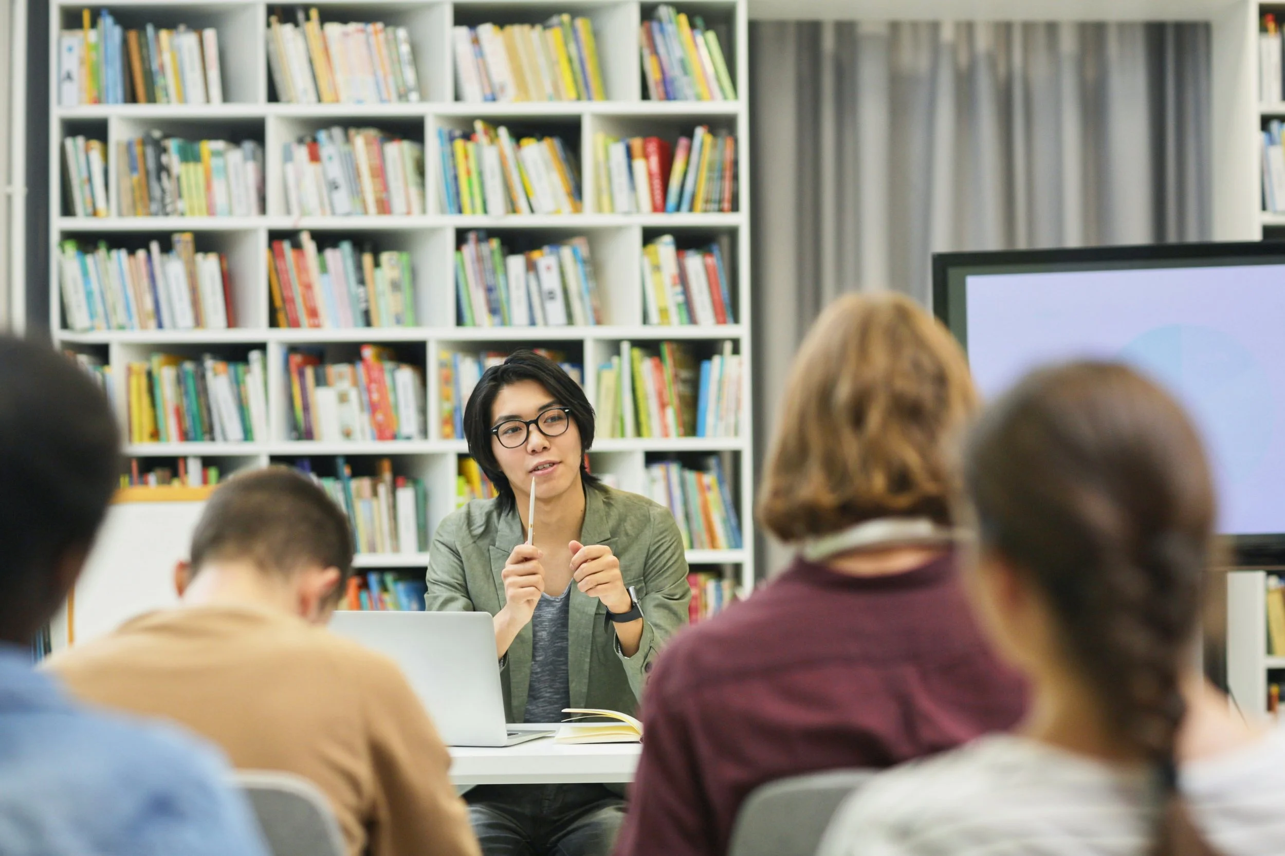 Asian person sitting in front a group of people with a laptop and pen in hand.  There are bookshelves in the background