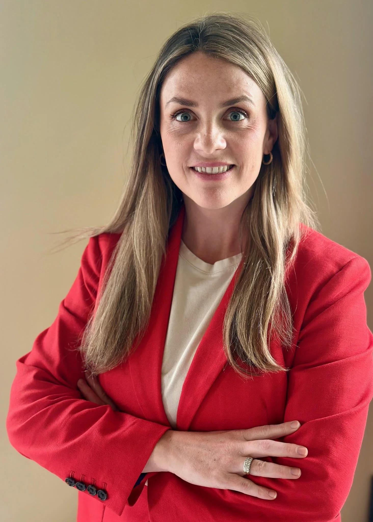 A woman with long blonde hair wearing a red blazer over a white top, smiling slightly, with her arms crossed.
