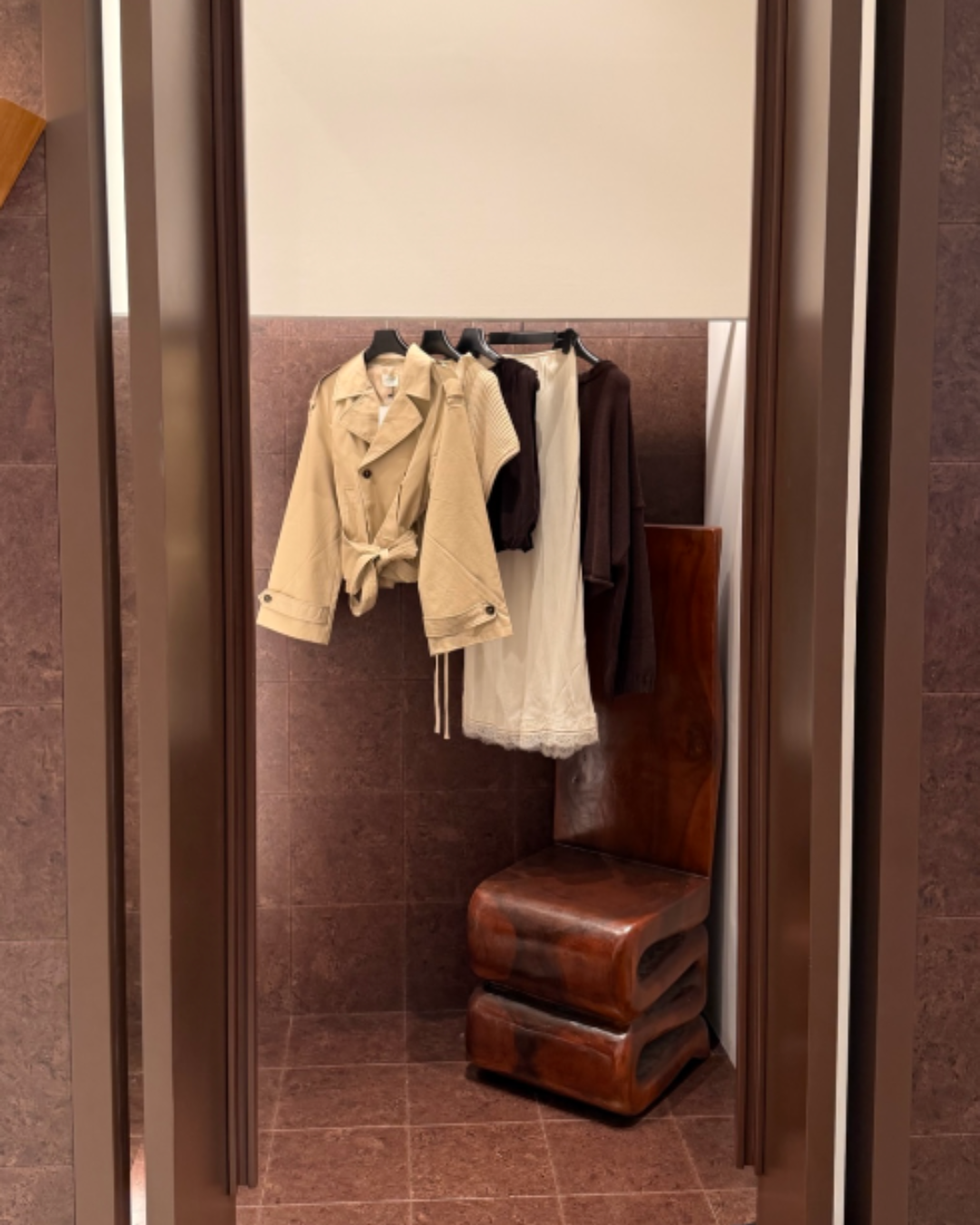 Clothes displayed in a fitting room with a wooden bench and beige, brown, and white clothing hung on a rack.