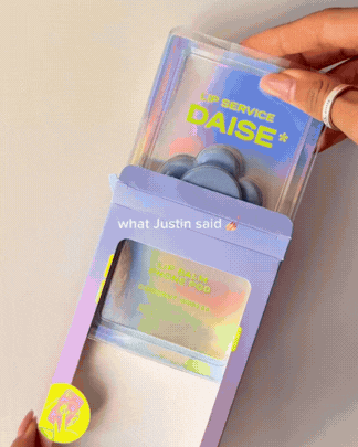 A hand holding a container of blue earbuds with a clear, holographic box, and a pink and purple package with a yellow sticker on the bottom left corner.