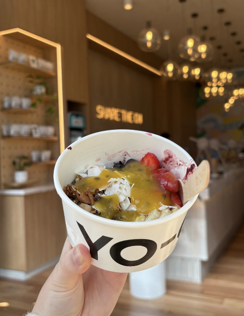 A hand holding a cup of frozen yogurt topped with strawberries, blueberries, blackberries, kiwi, syrup, and a cookie in a modern cafe setting with warm lighting and wooden decor.