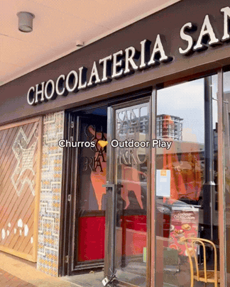 The storefront of a chocolate shop called 'Chocolateria San' with glass doors, a red chair outside, and reflections of buildings in the glass.
