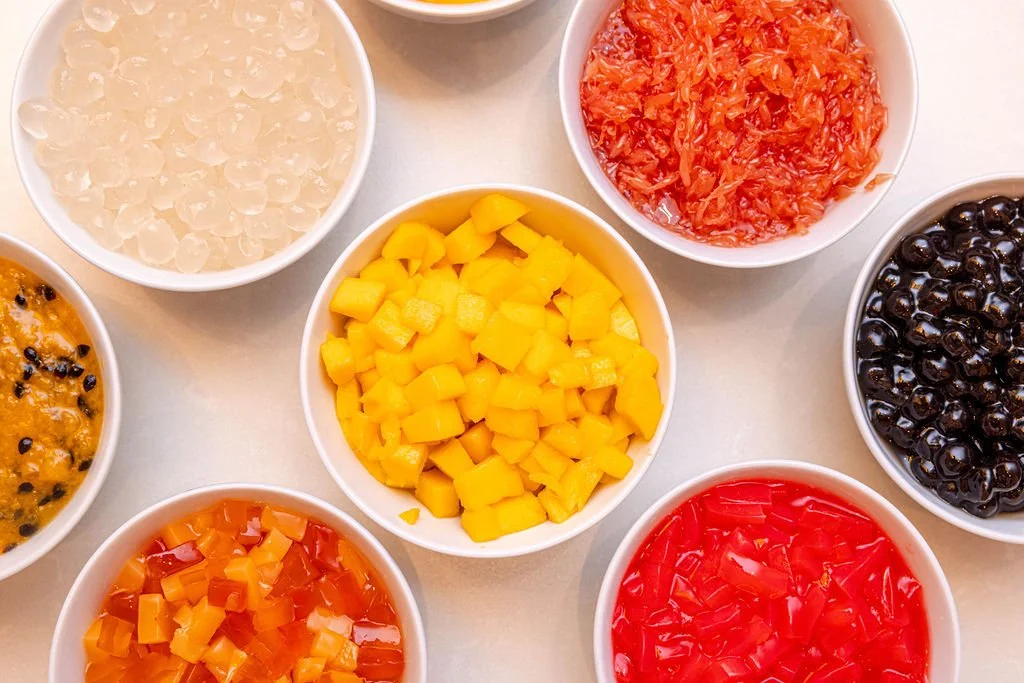 Eight small white bowls filled with different colorful chopped fruits and vegetables, including yellow mango, red tomato, black beans, and other diced produce, arranged on a white surface.