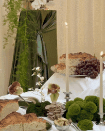 A table set for a meal with candles, bread, grapes, and green plants in a bright room with beige curtains.