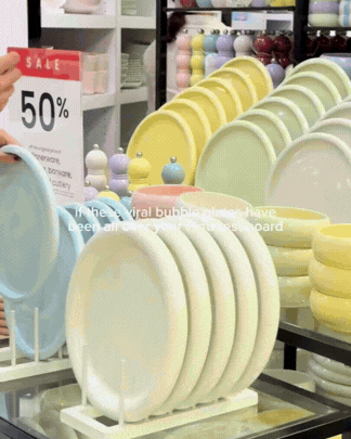 Display of yellow and white ceramic plates arranged on a rack in a store, with colorful wine glasses in the background and a red sale sign indicating 50% off.