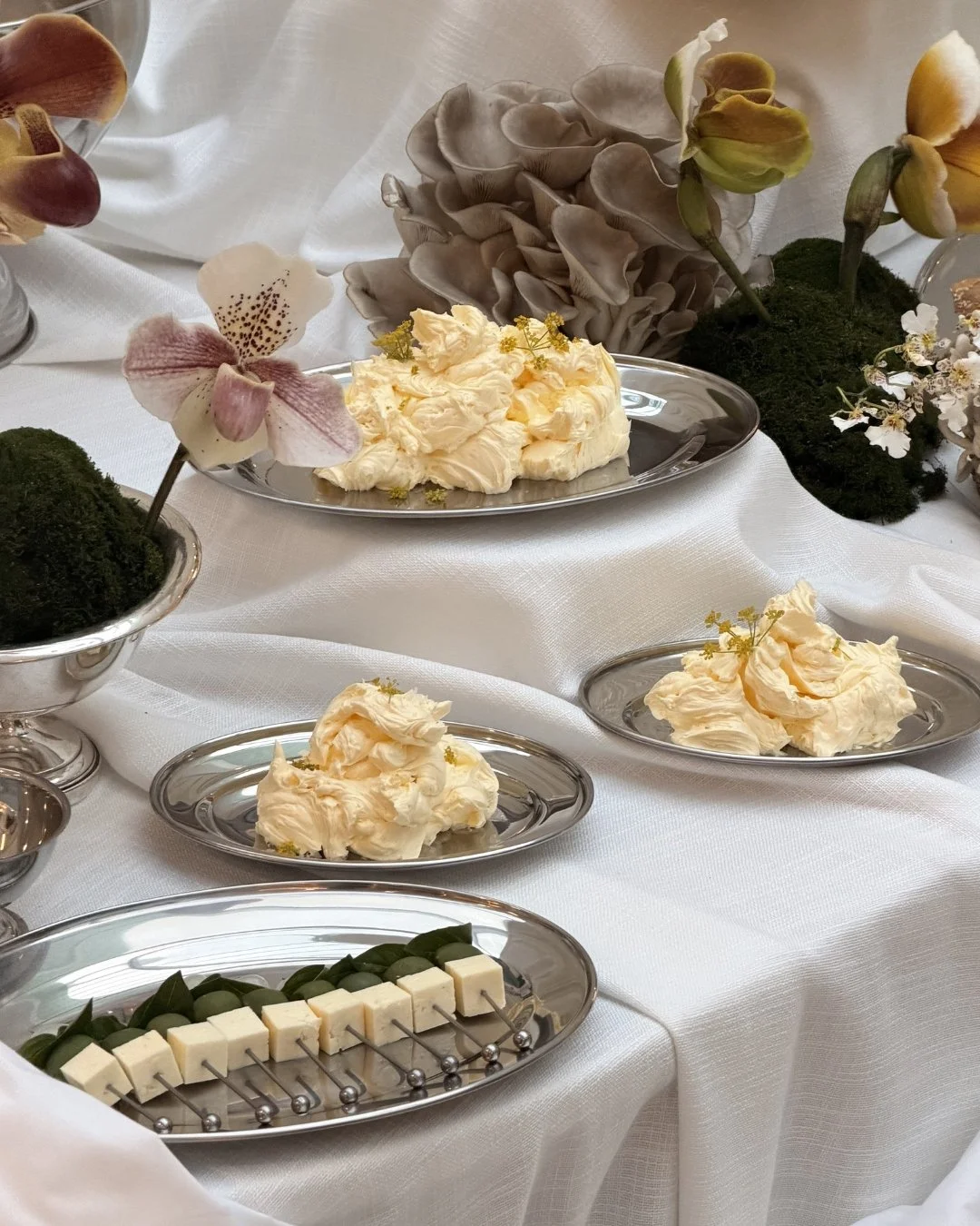 Elegant table setting with silver platters of whipped butter, cheese, and green olives, surrounded by decorative flowers and moss, on a white tablecloth.