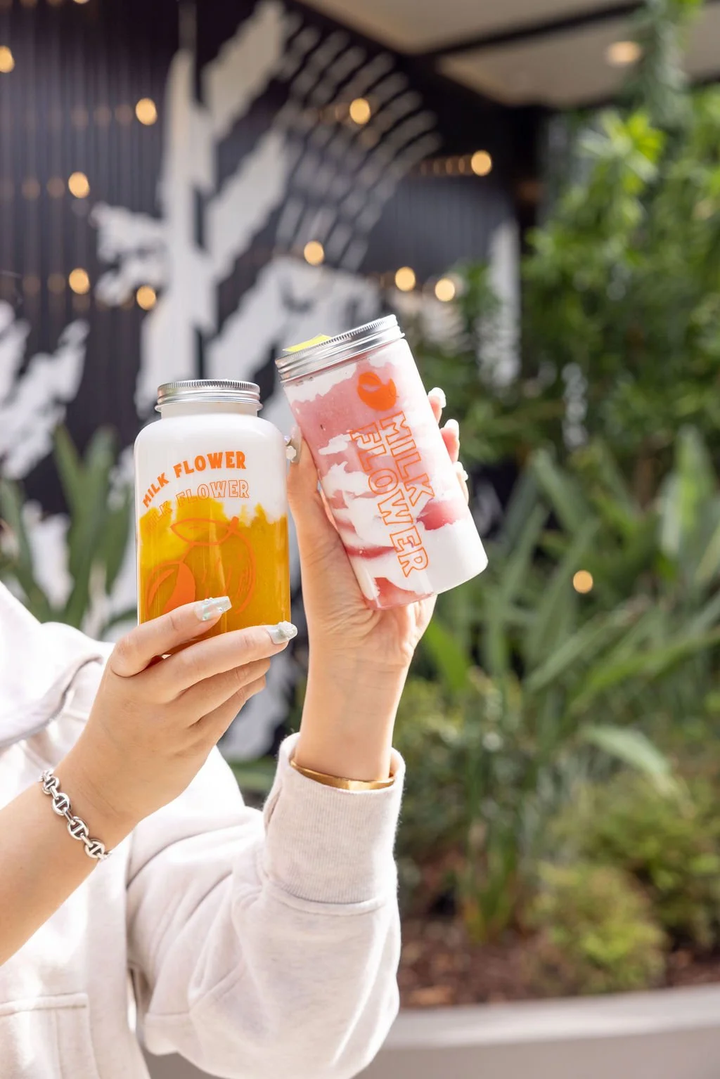 Two hands holding up beverages, one in a clear jar with an orange liquid and the other in a clear cup with a pink and white layered drink, against a backdrop of greenery and a decorative black and white wall.