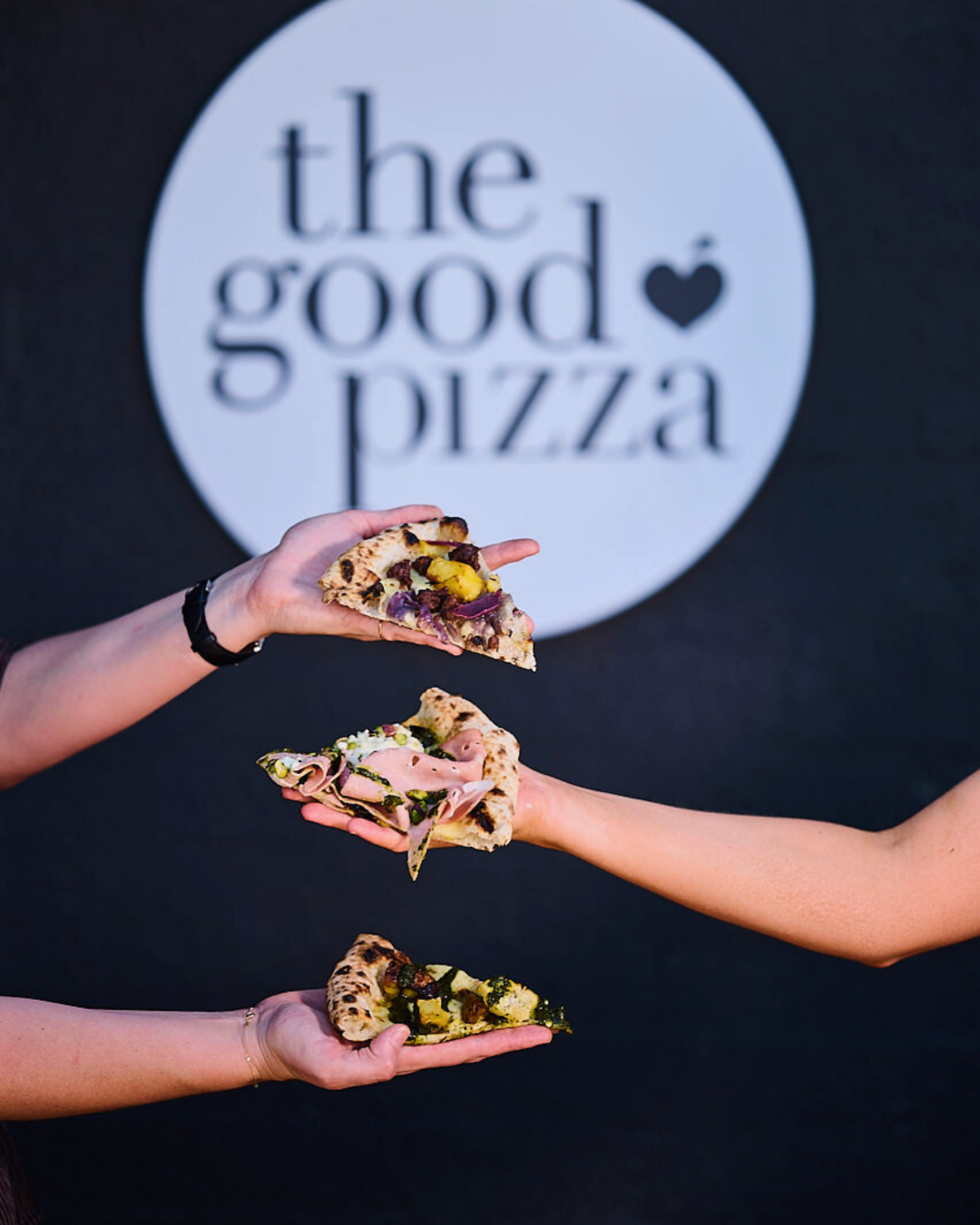 Three hands holding slices of pizza in front of a sign that reads 'the good pizza' with a heart symbol, against a dark background.