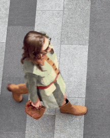 A woman walking on tiled floor, wearing glasses, a beige coat, brown boots, and carrying a purse