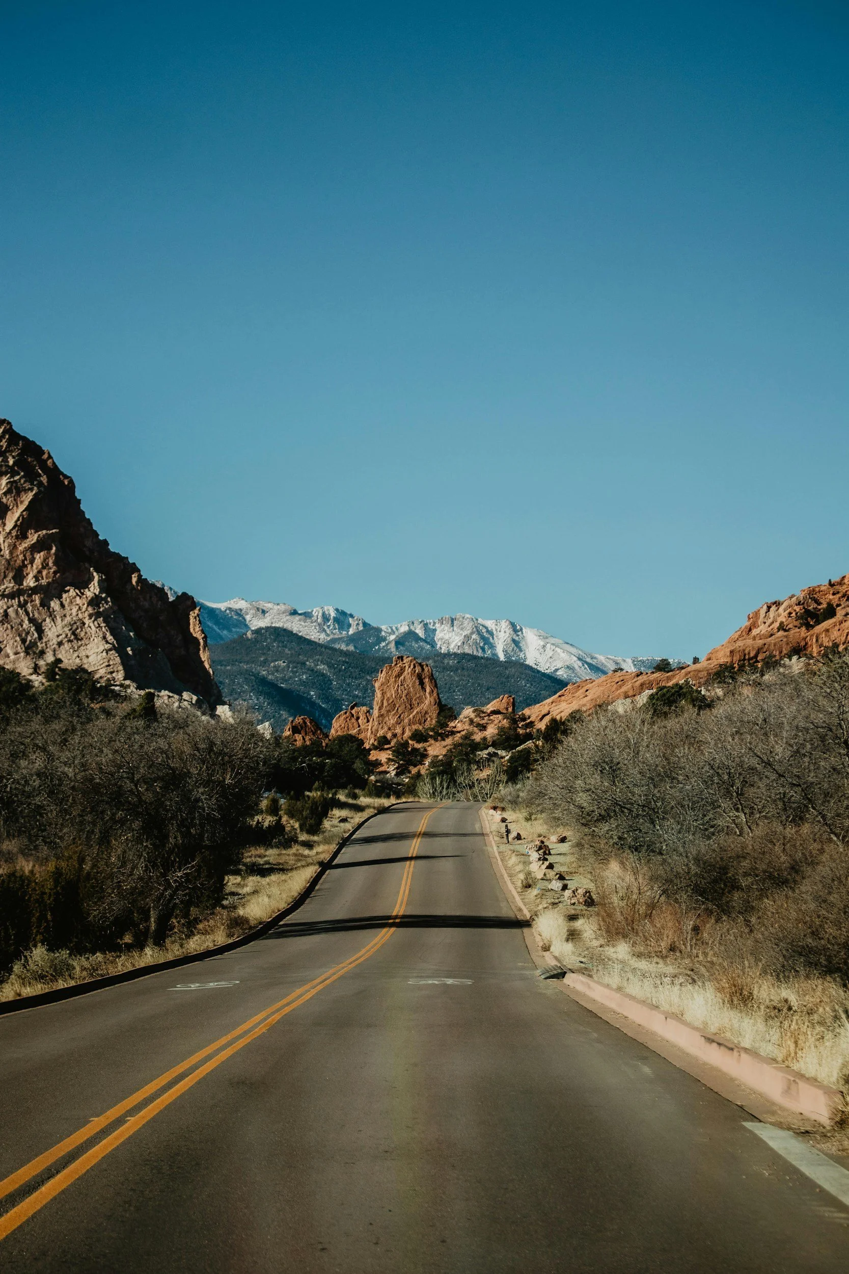A winding mountain road with double yellow lines, bordered by trees and rocks, leading towards snow-capped mountains in the distance under a clear blue sky.