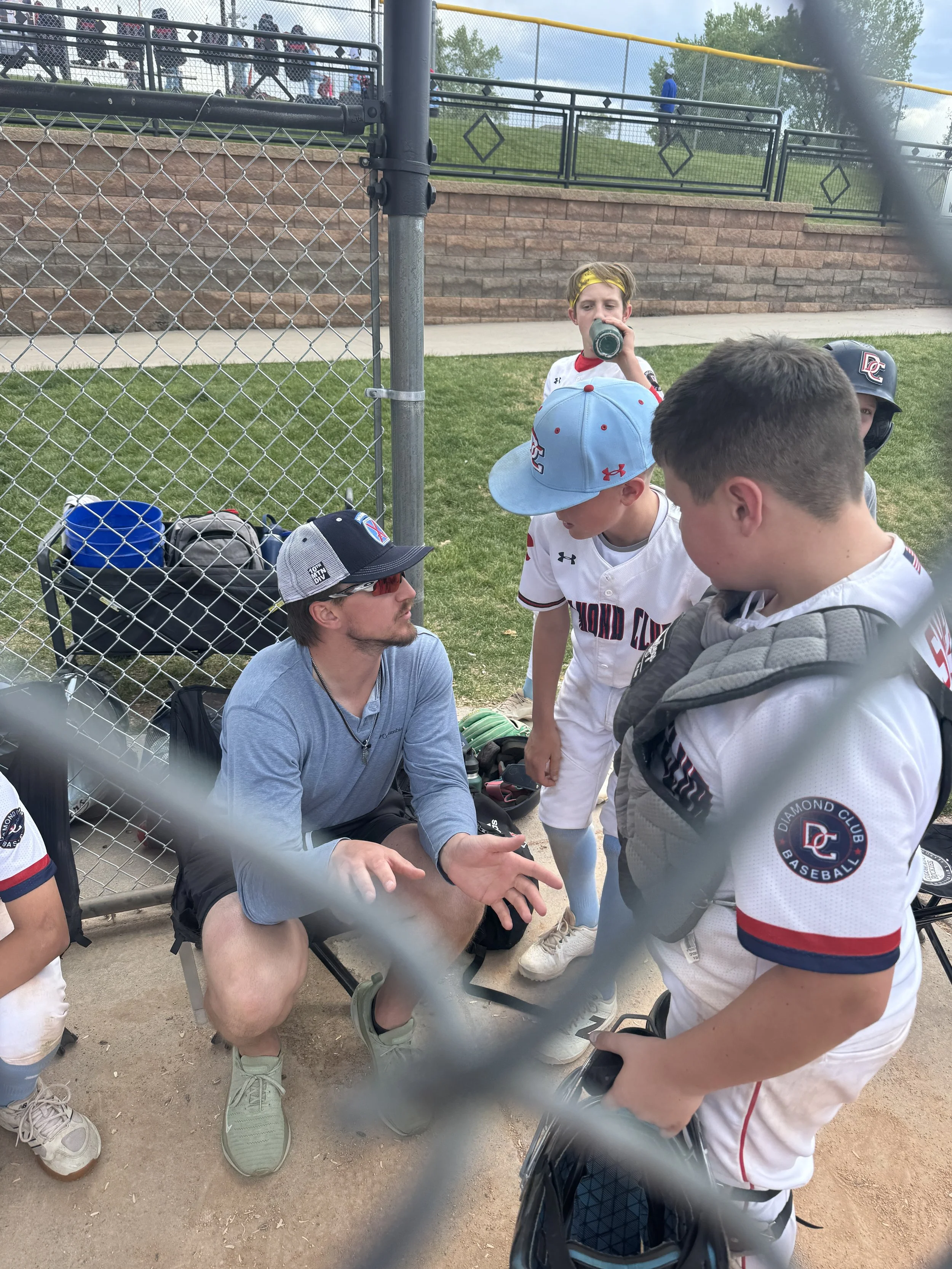 A group of young baseball players and a coach are gathered near a chain-link fence, having a discussion at a baseball field.