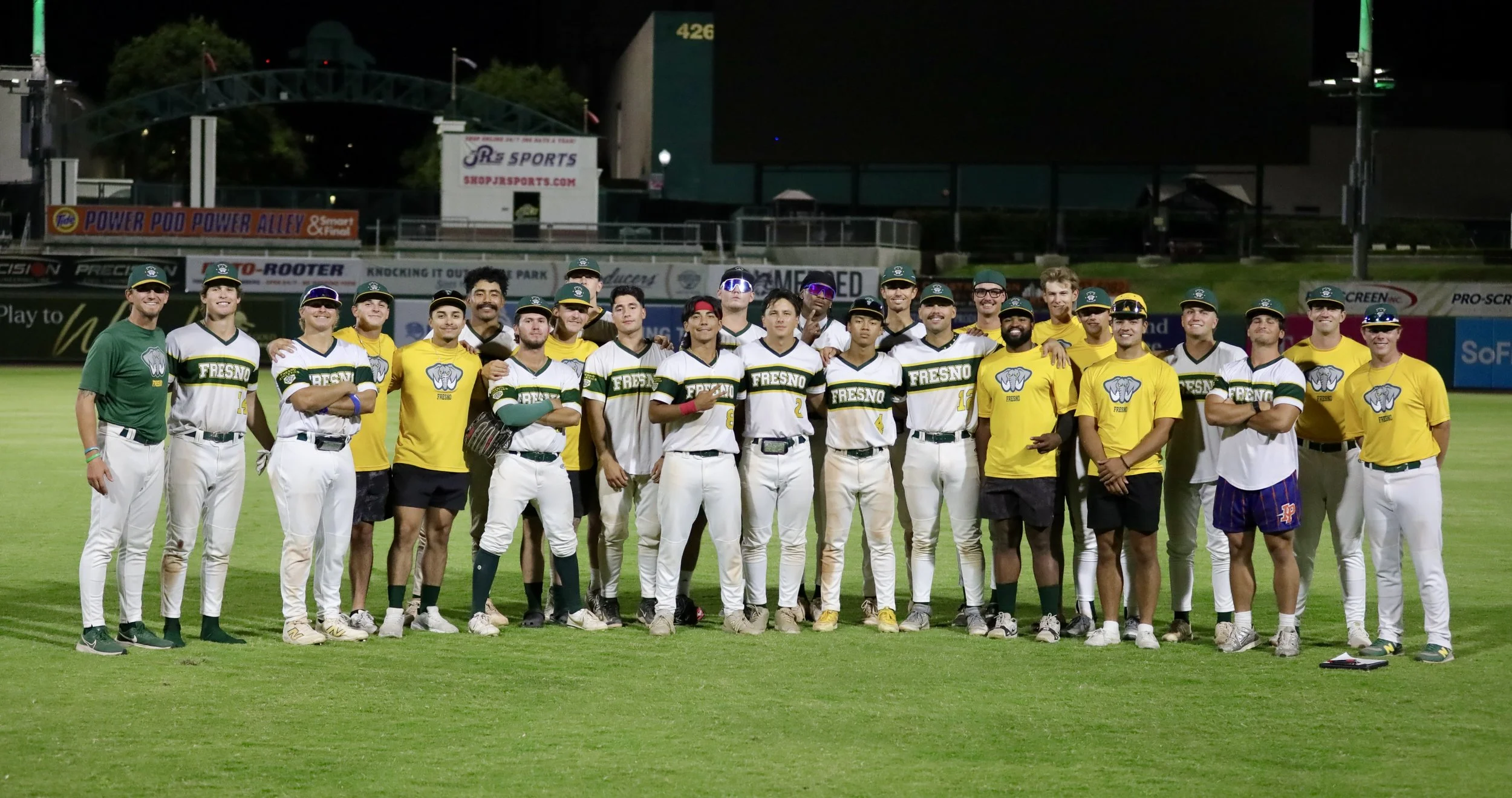 A group of baseball players and coaches standing together on a baseball field at night after a game or practice, with a stadium and advertisements in the background.