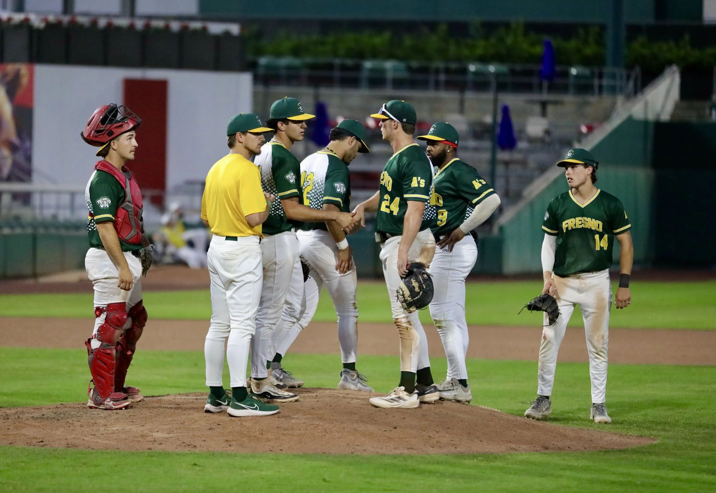 A group of baseball players, including a catcher in red gear and a pitcher in a green cap, stand on the field during a game. They are greeting and shaking hands with the opposing team. The scene is set in a baseball stadium with stands and advertisements in the background.
