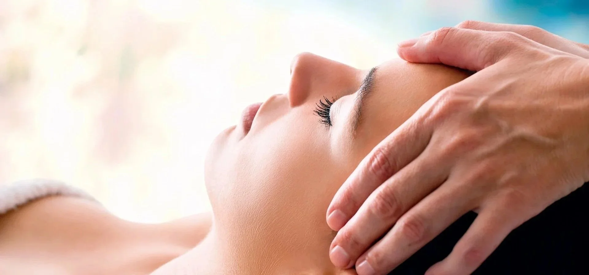 Close-up of a woman receiving a Reiki session, lying down with eyes closed, with hands on their forehead.